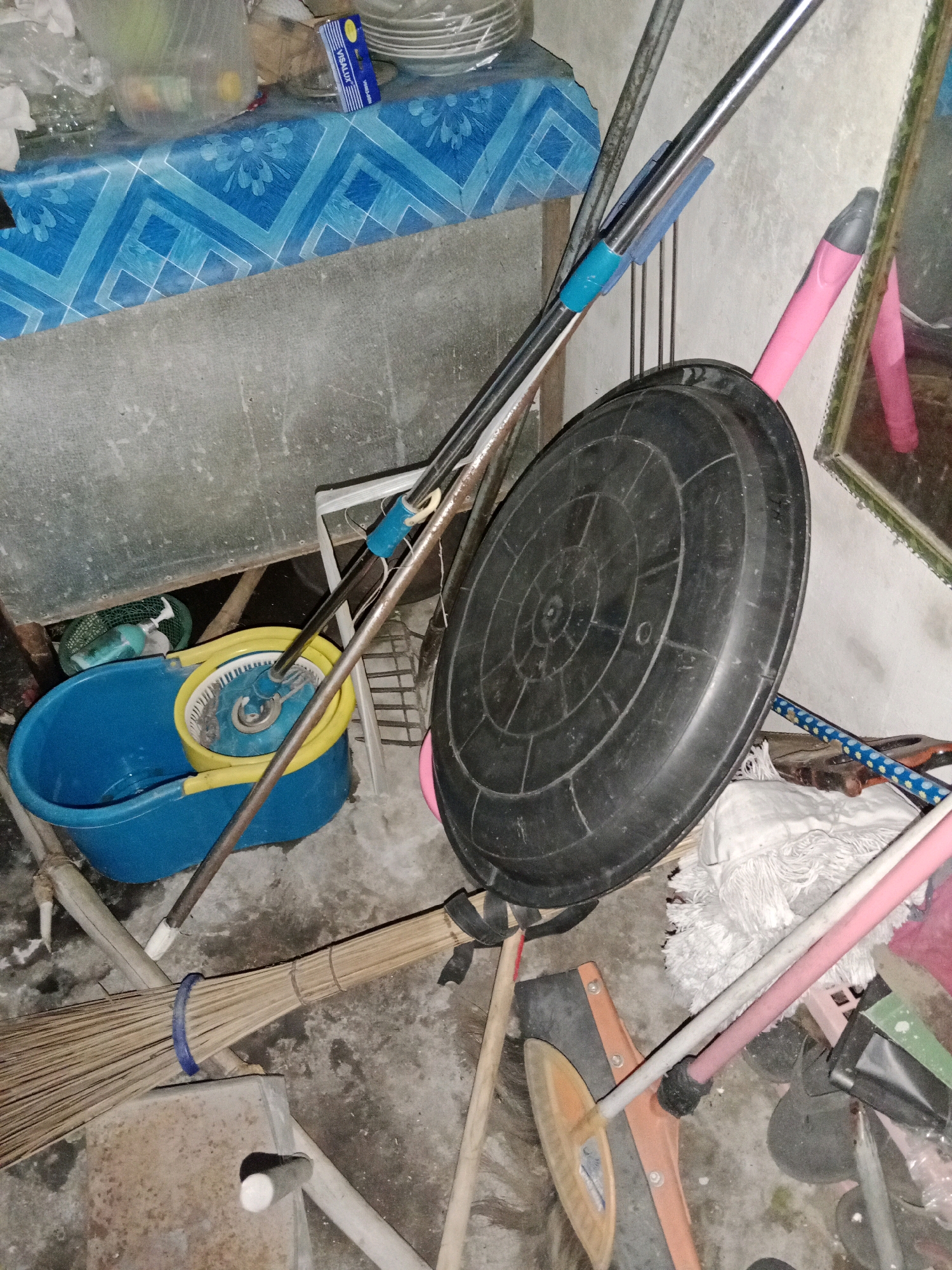 The image depicts a cluttered storage area, likely in a home or utility room in Ringinrejo, Indonesia. The scene is dominated by cleaning supplies, including a blue bucket with a yellow mop attachment, a broom with natural bristles and a blue tie, and several mops with white and pink handles. A large, black circular object, possibly a lid or a tray, is propped up against other items. Cleaning tools are leaning against a textured, light-colored wall. In the background, a table is covered with a blue and white patterned cloth, and a stack of white plates is visible. The floor is concrete and appears somewhat dusty. The overall impression is one of disarray and active use of the space for storing cleaning equipment.