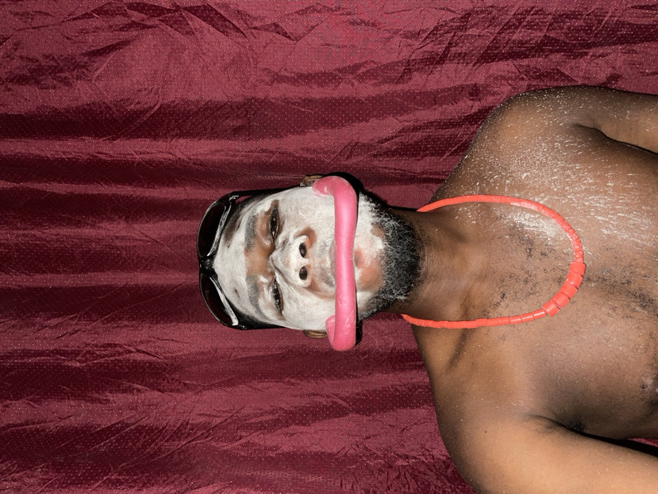 A man with sunglasses and a white face mask lies on a red, textured surface. A pink, hose-like object stretches across his face. He wears a segmented, orange necklace. His dark skin contrasts with the white mask and the surrounding red background.