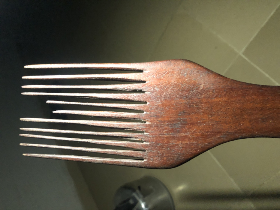 A wooden hair pick with many tines is angled across the frame. The tines are not all the same length; some are broken or shorter than others. The pick shows wood grain detail and an overall reddish-brown tone. A blurry background suggests a light surface with hints of a metallic object. The lighting emphasizes the texture of the wood.
