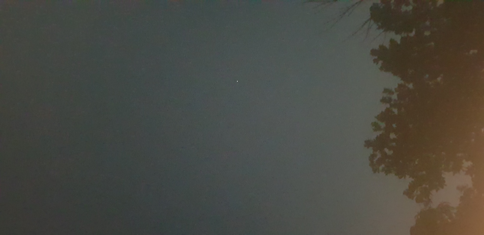 The image shows a dark, hazy sky with a single, bright point of light, likely a star or planet, visible in the upper center-right portion of the frame. The bottom portion of the image is filled with the silhouetted branches of trees, appearing dark against a faintly illuminated background. There are no people, animals, or vehicles visible in the image. The scene suggests an outdoor setting at night or during twilight, with some level of light pollution or atmospheric haze. The location in the city of Keboharan, Indonesia, indicates a likely urban or suburban environment.
