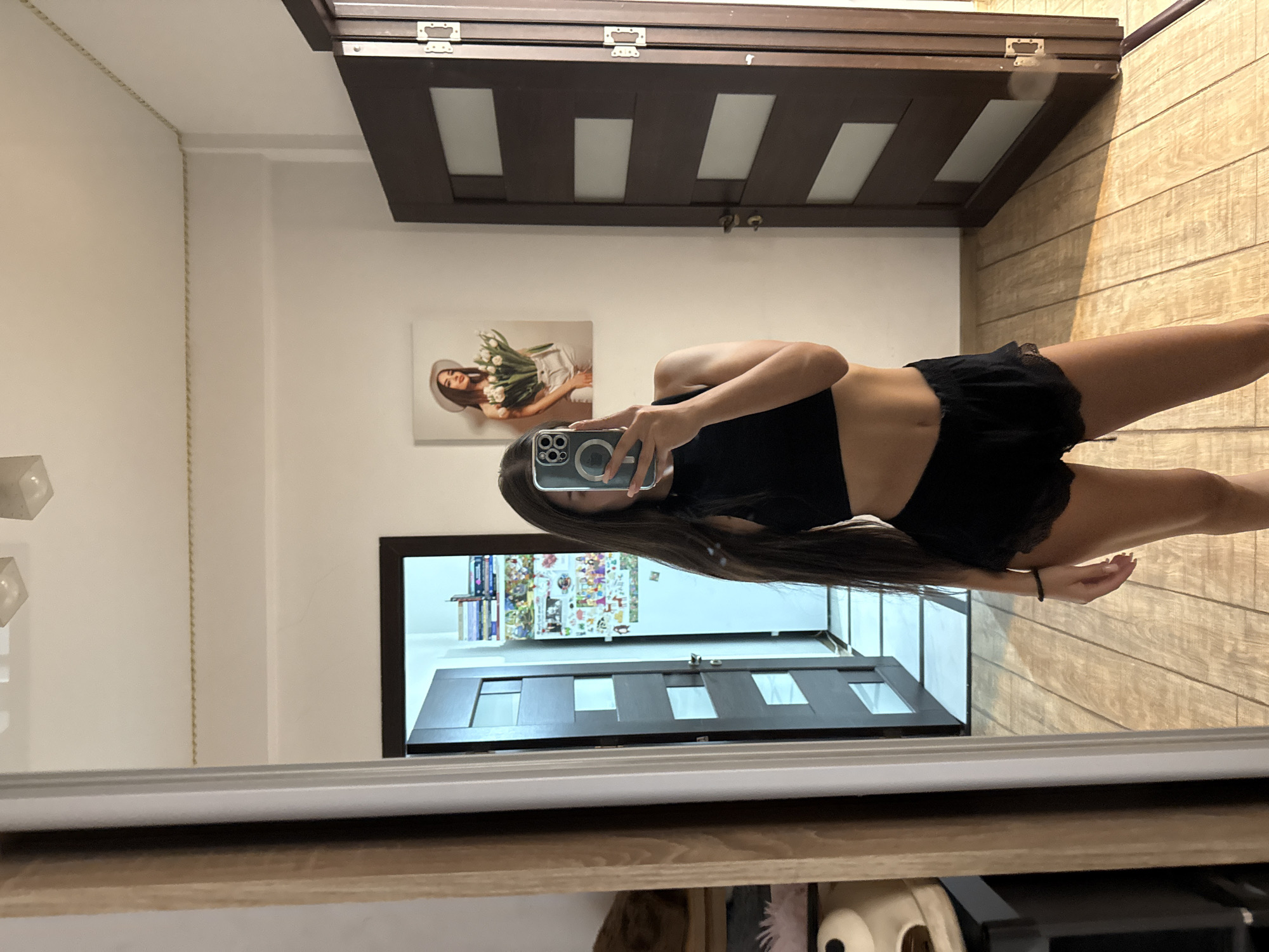 The image shows a young woman in black sleepwear taking a mirror selfie in what appears to be a bedroom or a dressing area. She is wearing a black crop top and black lace-trimmed shorts. Her long dark hair cascades down her back. In the reflection of the mirror, a painting of a woman with a hat and tulips is visible on the wall, along with a door with glass panels and a refrigerator decorated with magnets and photos. The room has wooden floors and white walls. The overall impression is a casual, personal moment captured in a domestic setting. The image does not contain information that would allow for its location to be identified as Chișinău, Moldova.