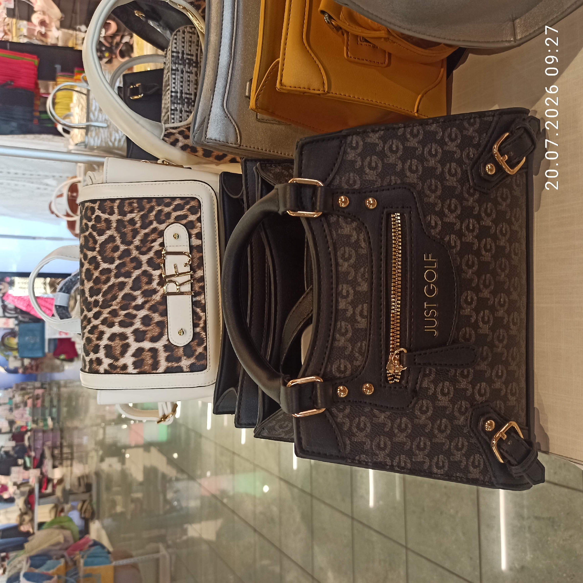 A collection of handbags is displayed on a shelf in a retail store. The foreground features a dark brown handbag with a repeating "JG" logo pattern, a gold zipper, and a white label that reads "JUST GOLF." Above it, a white handbag with a leopard print front panel and gold hardware is visible. Further back, other handbags in various colors and textures can be seen, including a metallic silver one and a mustard yellow one. The store has a polished floor reflecting the lights and displays. The date stamp "20.07.2026 09:27" is overlaid on the bottom right of the image. No people are visible in the image, and no specific location in the city of Bari, Italy, can be identified from the visual evidence.