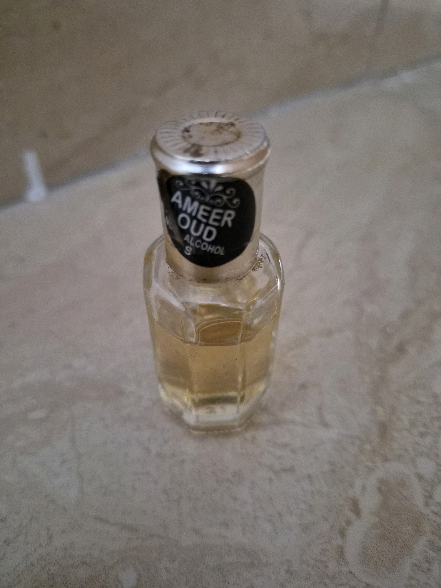 A small, faceted glass bottle containing a light yellow liquid sits on a mottled, beige tiled surface. The bottle has a silver, ridged cap with a black label that reads "AMEER OUD ALCOHOL S". The background is a blurred, beige wall with a faint white vertical shape. The scene is indoors, likely a bathroom or tiled room, in Srimahi, Indonesia.