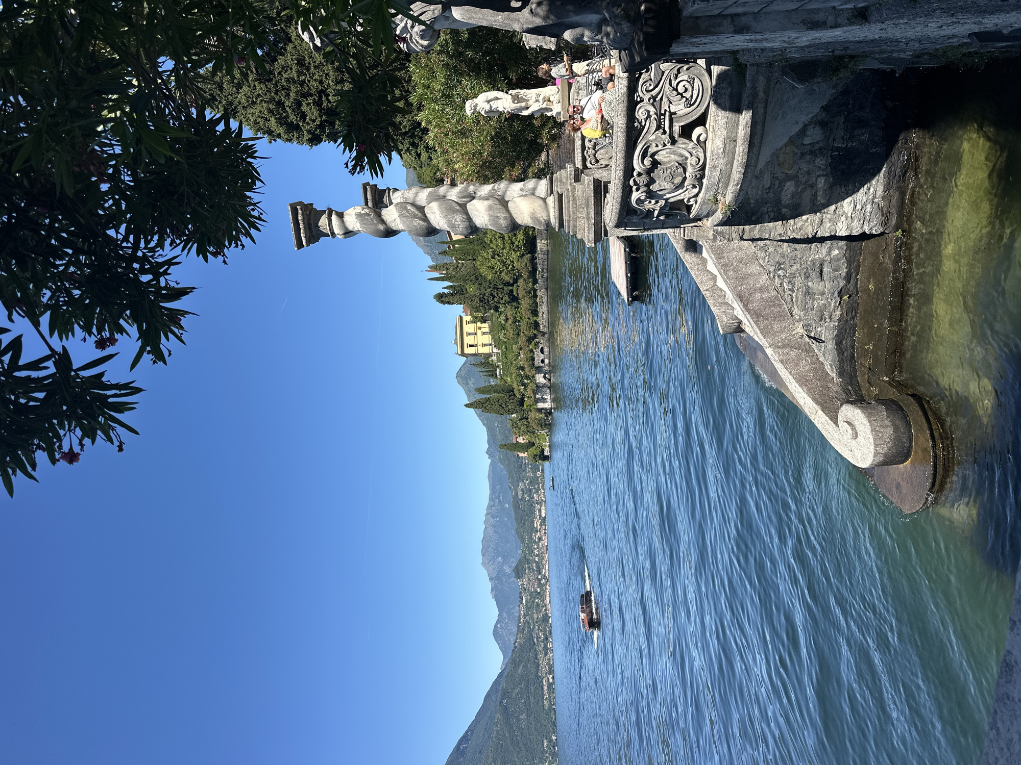 This is a full-frame, eye-level, outdoor, bright daylight shot of a serene lakeside scene, likely in Varenna, Italy, given the context of the image and common tourist destinations.
In the foreground, a stone balustrade, featuring intricate carvings and ornate scrollwork, forms the right edge of the frame. To the left of the balustrade are stone steps leading down to the water. A large, twisted stone column stands prominently behind the balustrade, partially obscured by lush green foliage overhead. To the right of the column, a white statue of a male figure stands on a pedestal, and two women are seated on a bench nearby. The water of a large lake, likely Lake Como, dominates the left side of the image. The water is a vibrant blue, with gentle ripples. A small boat with a red top is moving away from the camera, leaving a wake.
In the middle ground, across the lake, are the lush green hillsides dotted with buildings. A prominent yellow building is visible on the hillside. The landscape is characterized by rolling hills, lined with cypress trees and other greenery.
The background consists of a clear, bright blue sky. Distant mountains, with a haze, are visible on the horizon, framing the lake and the landscape on the opposite shore. The overall atmosphere is peaceful and picturesque, evoking a sense of tranquility and natural beauty.