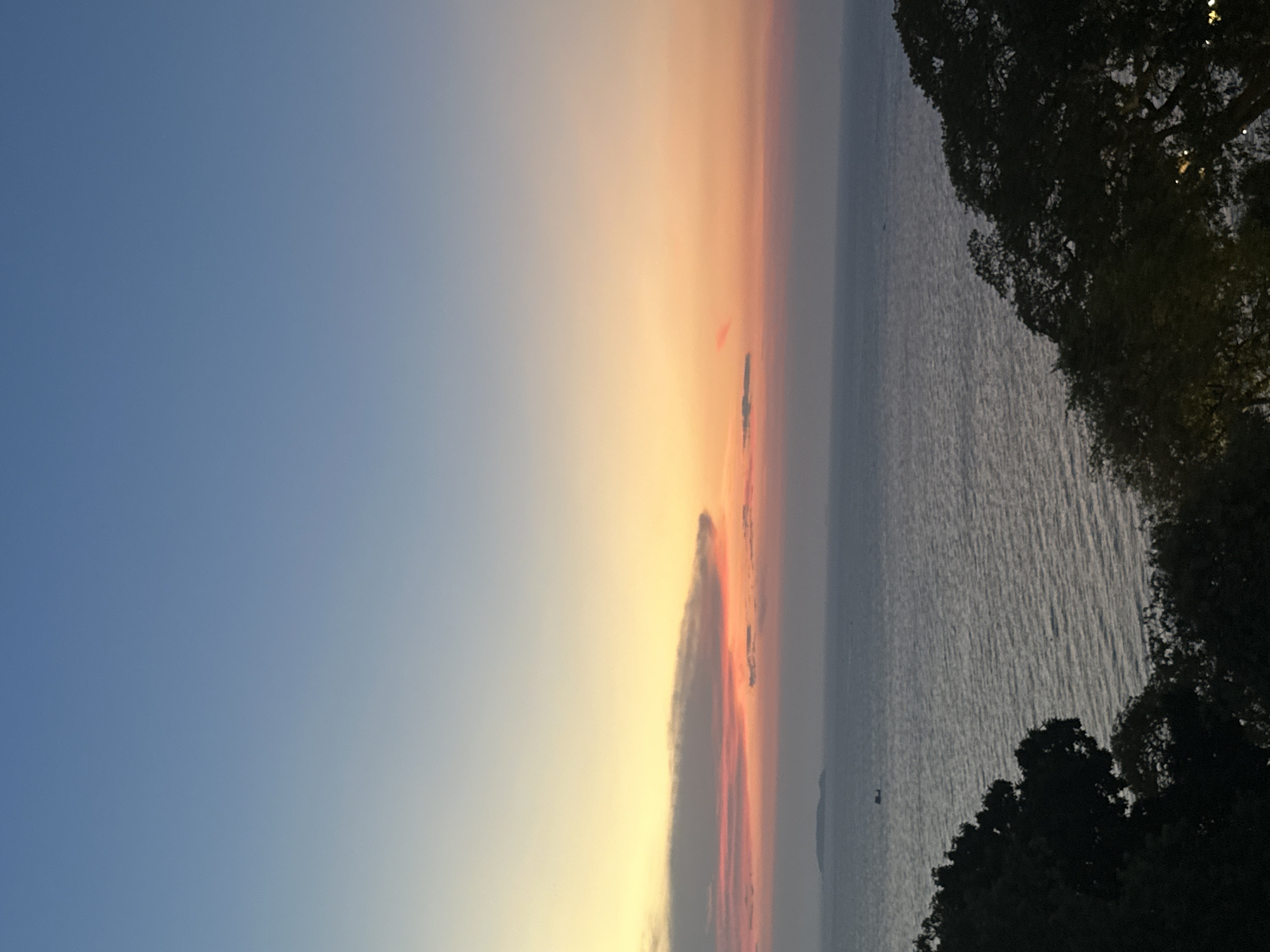 The image depicts a sunset over the ocean, likely in Pattaya City, Thailand, based on the landscape and geographical features. The sky transitions from a clear blue on the left to shades of orange, pink, and yellow near the horizon. A layer of clouds near the horizon displays shades of grey and pink. The ocean is visible to the right, exhibiting a rippled surface. Silhouetted trees line the right edge of the frame. A small boat is visible on the ocean in the middle of the frame.