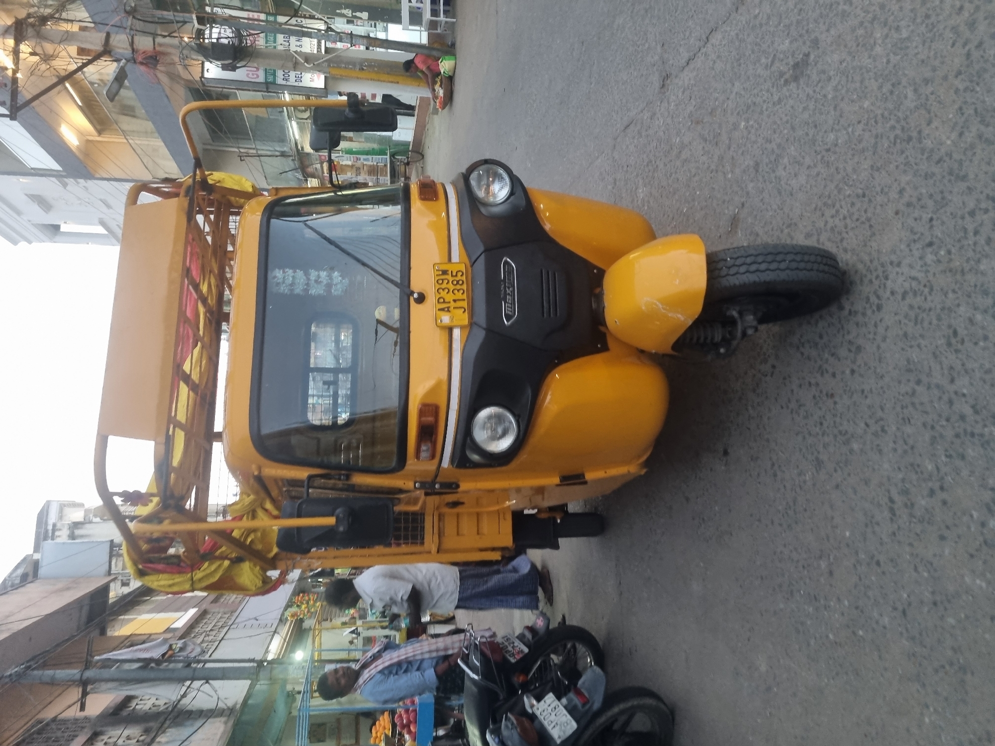 The image captures a yellow auto-rickshaw in Puttaparthi, India. The auto-rickshaw, bearing the license plate "AP39W J1385," is parked on a paved road. Two men are visible, one standing near the auto-rickshaw wearing a white shirt and dark trousers, and another man in a blue shirt examining a motorcycle. The street scene includes buildings, shops, and street vendors. The buildings have signs in Telugu and English, notably including a sign for "ROCS LAB & H". The overall environment suggests a typical street scene in Puttaparthi, with local businesses and transportation.