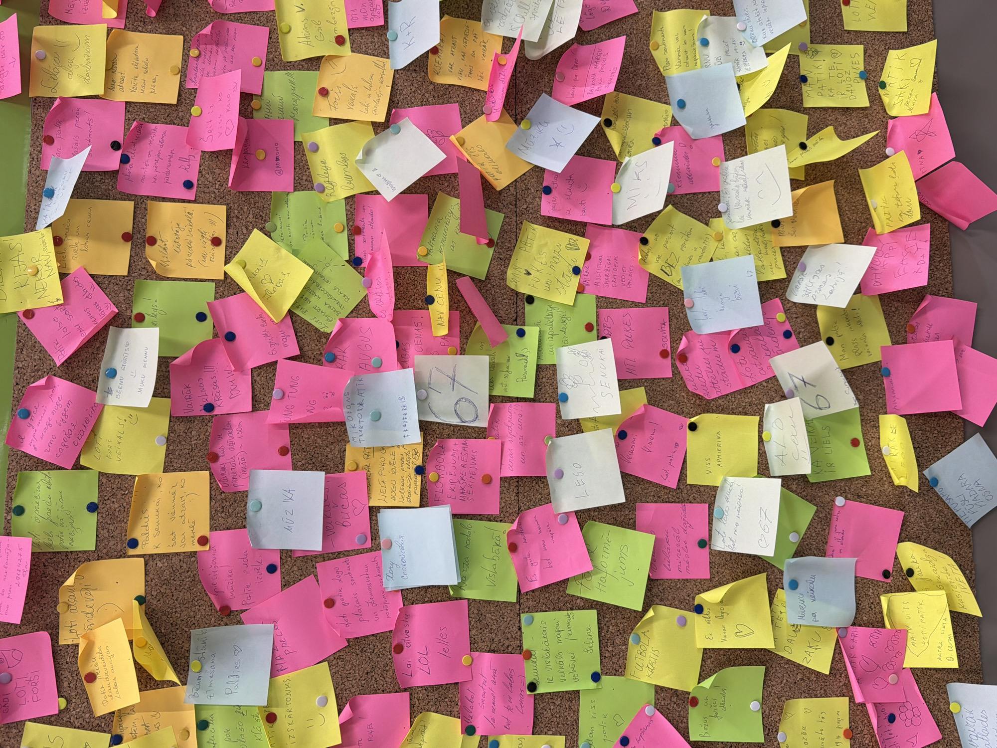 The image shows a corkboard densely covered with colorful sticky notes, predominantly in pink, yellow, and green. The notes are attached with various colored pushpins. Most of the notes appear to have handwritten text in Latvian. The environment is indoors. Many notes express positive feedback or requests. Key terms are visible: LEGO, SmartNet, Depo, Senukai. The board may be located in Riga, Latvia.