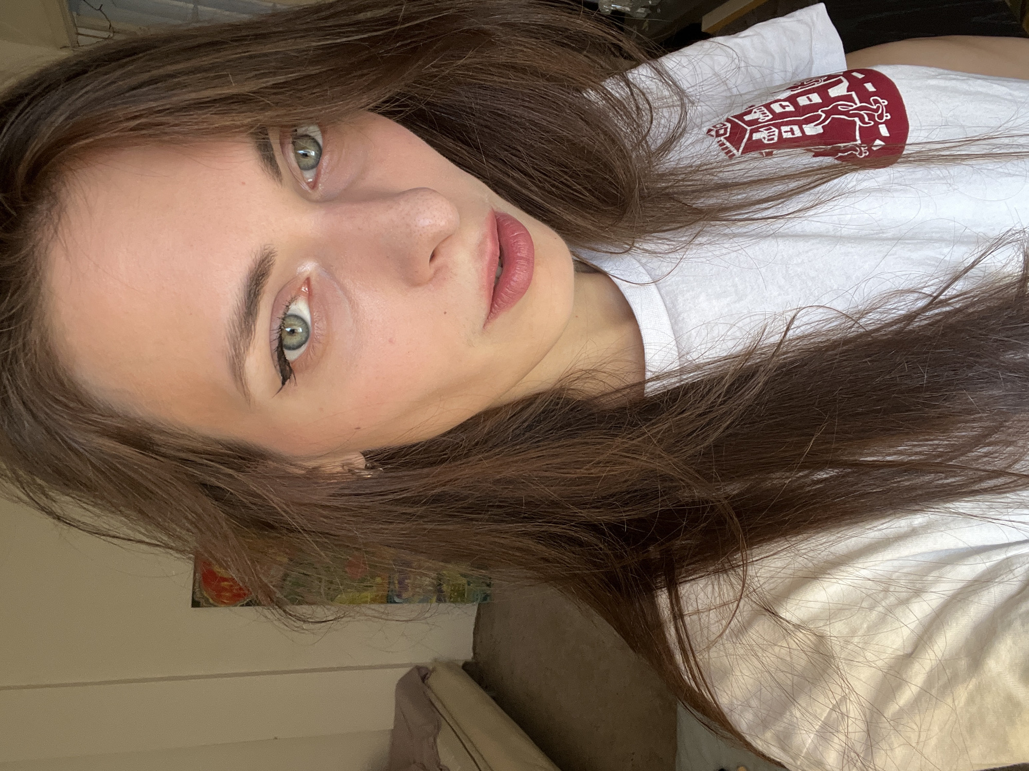 A woman with brown hair and green eyes looks at the camera. She wears a white t-shirt with a maroon design on the left sleeve. The background shows part of a white wall with a small painting visible, and a portion of a gray sofa with bedding. The location is not identifiable as Krasnoyarsk, Russia, based on the image alone.