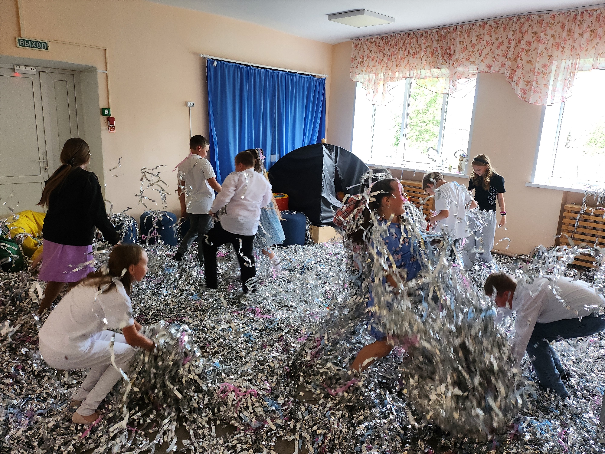 Location: Krasnogorskoye, Russia, interior of a room.
Scene: A group of children are actively playing with silver and pink confetti scattered across the floor. The children are dressed in a variety of clothing, including white shirts, dresses, and leggings. Some children are standing, walking, and bending over, gathering handfuls of confetti. The room appears to be a classroom or playroom, with a window covered by floral-patterned curtains. Blue curtains hang in the background. A sign with the word "ВЫХОД" (EXIT) in Russian is located near the door in the background. A small black tent is visible near the center.