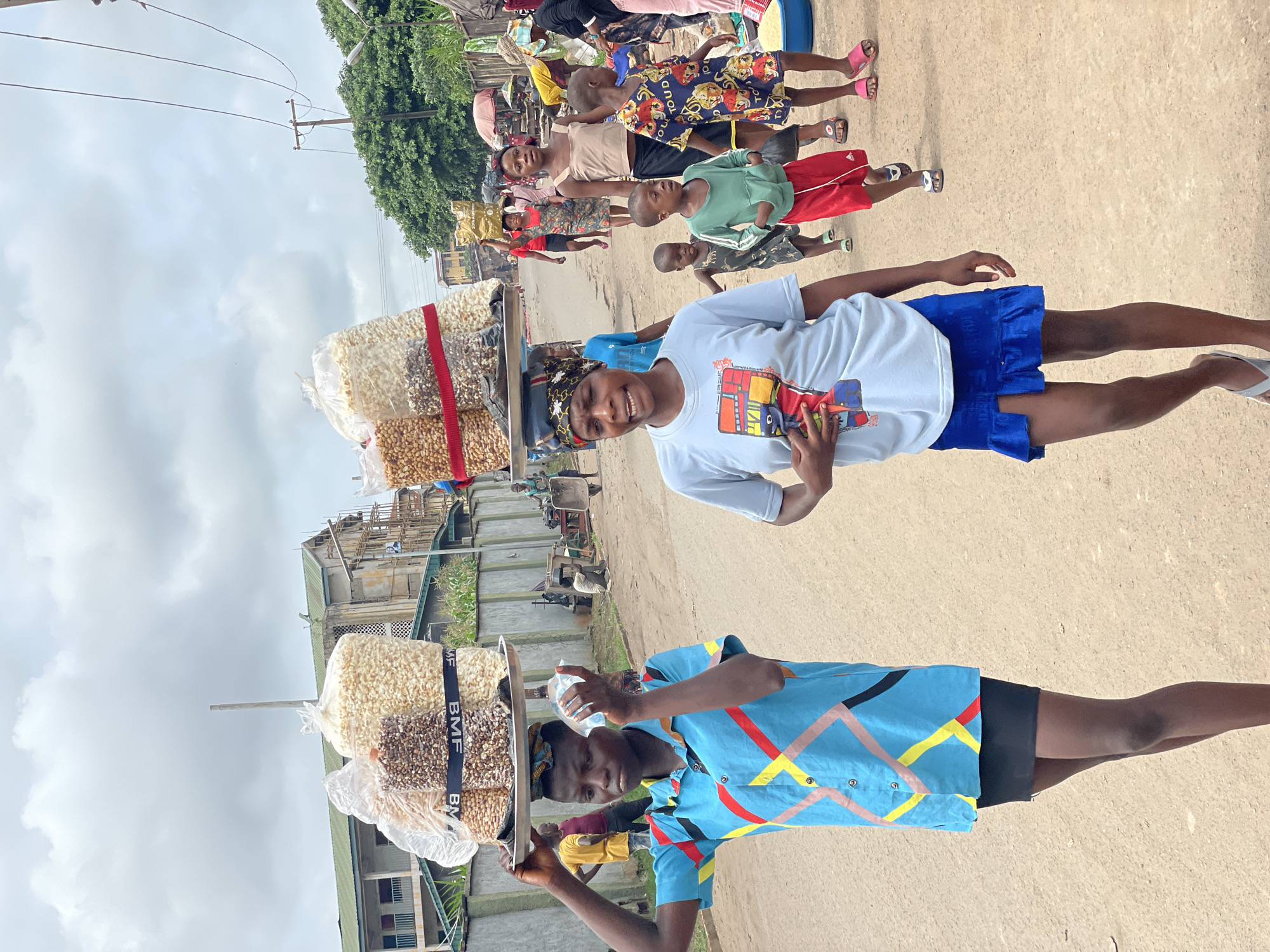 The image captures a vibrant outdoor scene on an unpaved road under a bright, partly cloudy sky. Two young individuals are prominently featured in the foreground, each balancing a tray of goods on their head, likely operating as street vendors.
The person on the left, smiling and looking towards the viewer, wears a white t-shirt adorned with a colorful graphic depicting a bus or truck, blue shorts, and white flip-flops. On their head, a flat tray holds several clear plastic bags filled with white popcorn and smaller, darker kernels, possibly peanuts or nuts. These bags are secured by black straps repeatedly printed with "BMF".
To their right, another young person, also looking towards the camera, sports a light blue button-up shirt featuring bold geometric patterns in yellow, red, and black, worn over black shorts. They hold a small clear plastic bottle in one hand while similarly balancing a tray of bagged popcorn and nuts on their head, also secured by "BMF" branded straps.
In the mid-ground and background to the left, a group of children of varying ages are visible, some standing, some possibly walking, dressed in casual attire including a patterned dress and a green shirt with red shorts. The overall setting appears to be a busy community area or market, with simple buildings, utility poles with overhead wires, and green foliage lining the dirt road. The time of day appears to be daylight, with the light suggesting late morning or early afternoon. The direct gaze and smiles of the foreground subjects convey a friendly interaction with the camera.