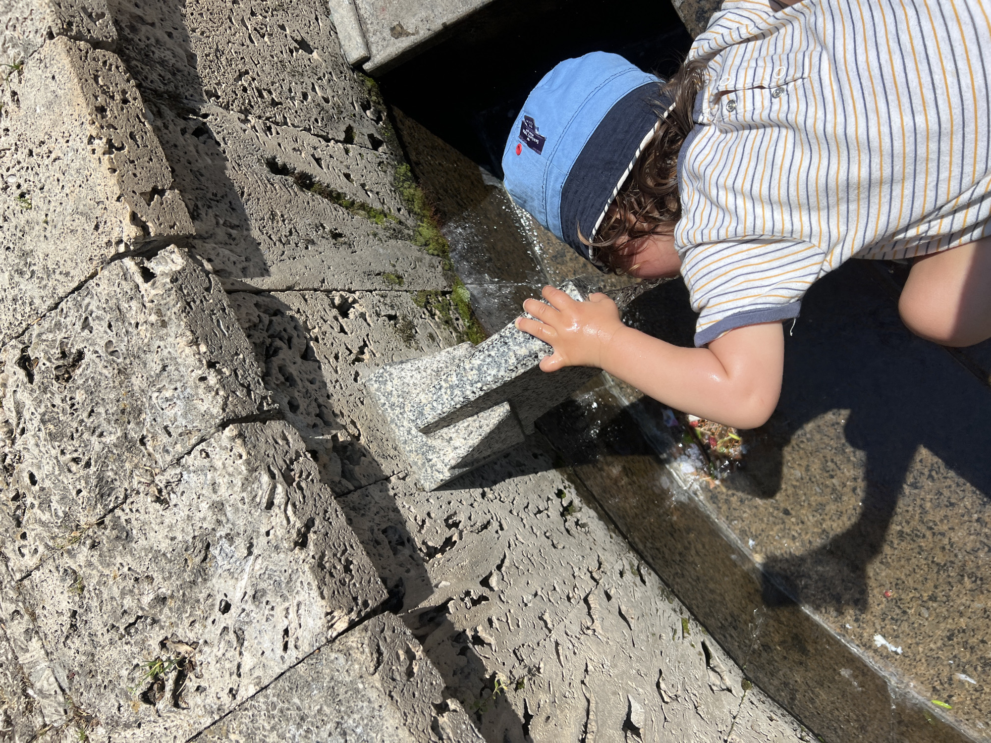 A young child wearing a blue and white bucket hat and a striped t-shirt is leaning over a stone fountain, playing with the water. The child's hand is visible, touching a small stone spout from which water is flowing. The fountain is constructed of rough, light-colored stone blocks. The scene appears to be outdoors in sunlight, casting shadows on the stone. Small plants and moss are growing in crevices of the stone. The water in the fountain basin is clear with some debris visible at the edge. The location is the Ivan Vazov National Theatre in Sofia, Bulgaria, specifically the fountain in front of the theatre.