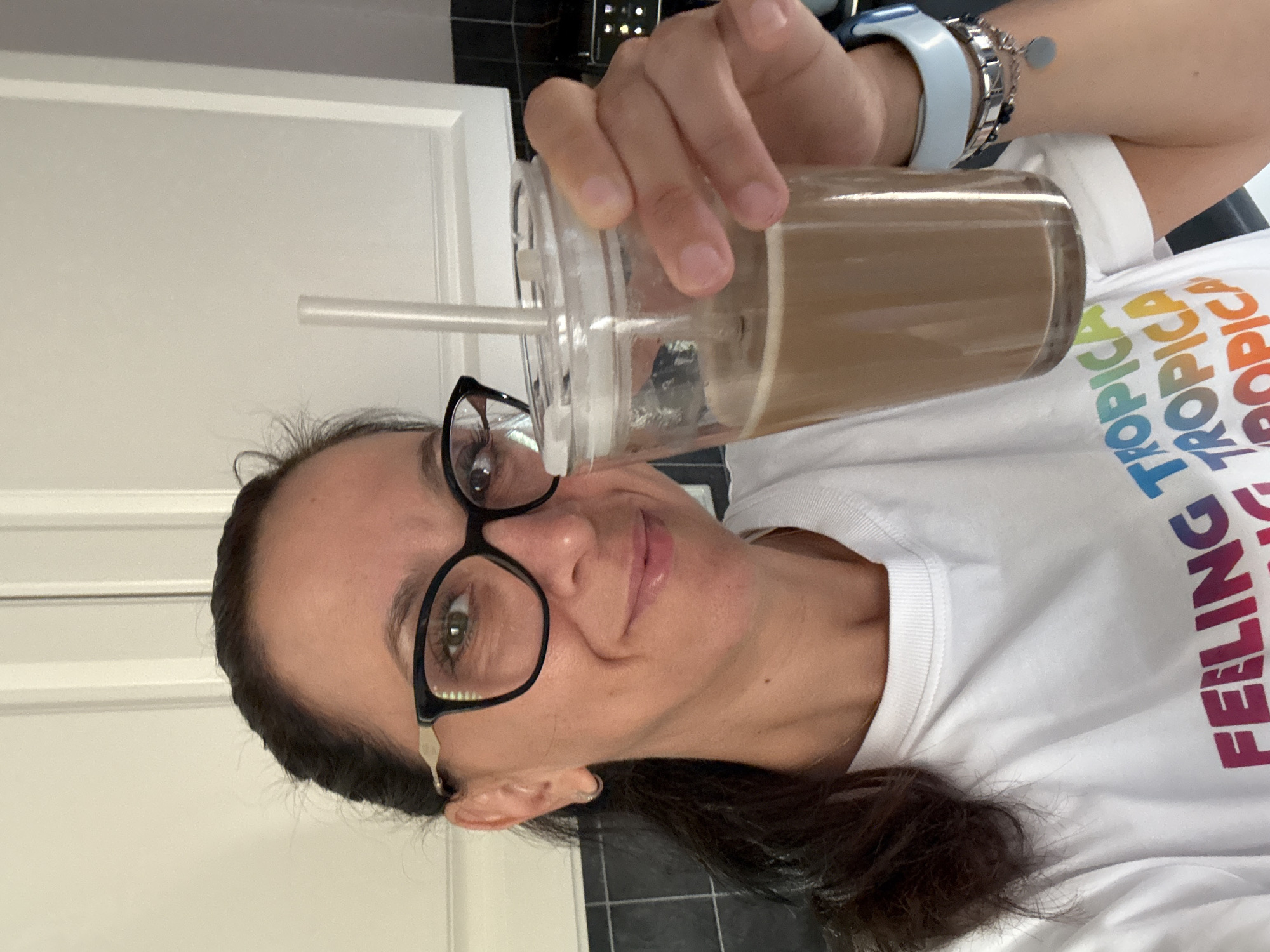 A woman with dark hair, wearing black-rimmed glasses and a white t-shirt, holds a clear plastic cup with a straw filled with a brown beverage. The t-shirt has colorful text on it that reads "FEELING TROPICA TROPICA TROPICA TROPICA". She is wearing a light blue smartwatch and several bracelets on her left wrist. The background shows white kitchen cabinets and a black tiled backsplash. The scene is indoors, likely in a kitchen. The location is Marum, Netherlands.