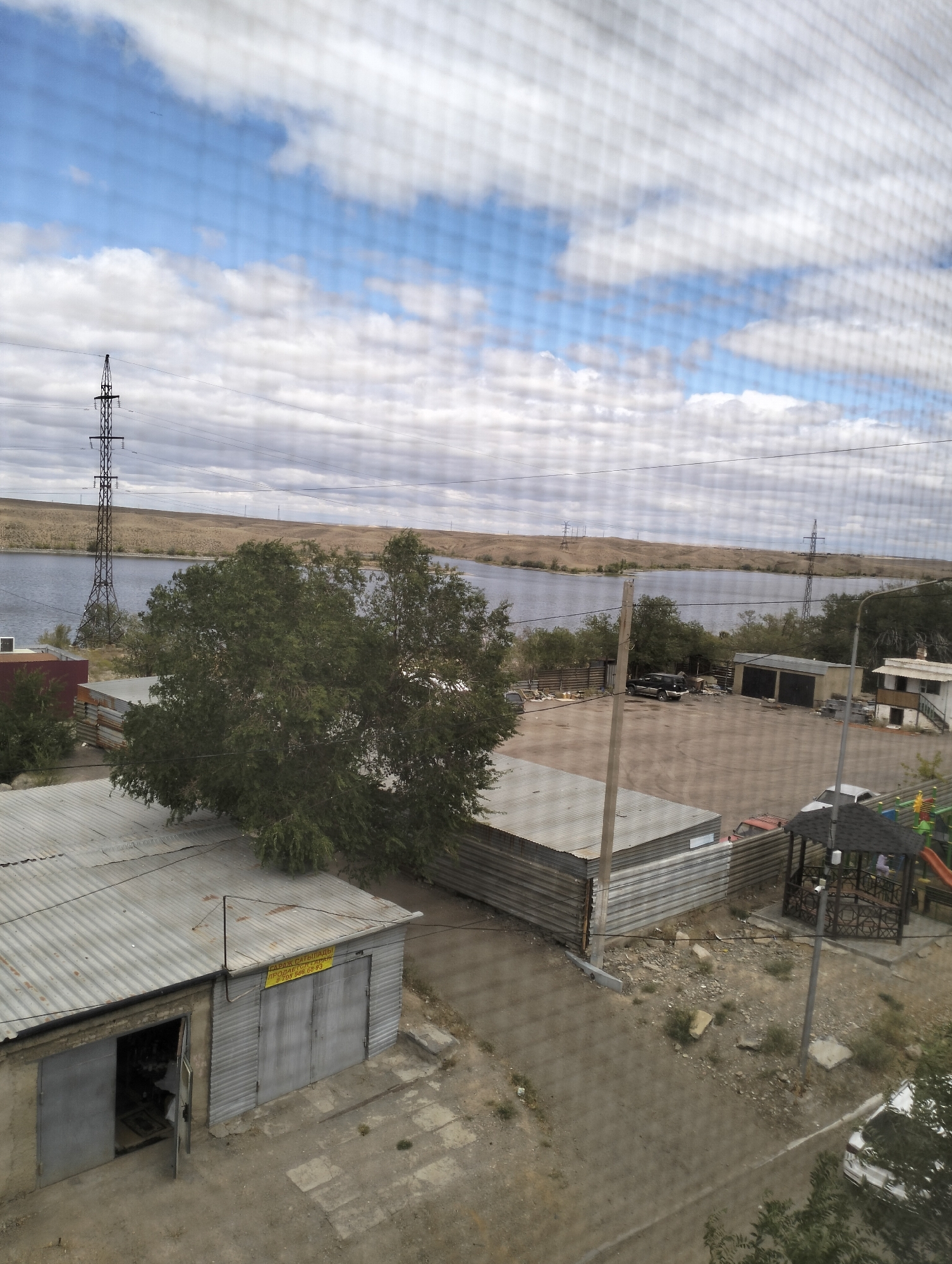 The image depicts a view of a body of water, likely a reservoir or river, with a barren, hilly landscape in the background. In the foreground, there are several industrial-style buildings with corrugated metal roofs, appearing to be garages or storage units. A large tree with dense green foliage is positioned between the buildings and the water. Several power lines and transmission towers are visible across the landscape. The scene is viewed through a mesh screen, indicating it was taken from inside a building or through a window with a screen. The presence of a children's playground with slides and a gazebo in the middle-right of the image suggests a residential or public area adjacent to the industrial structures. Cars are parked in the lot. The overall impression is one of a functional, possibly somewhat utilitarian area in a region with a dry climate, characteristic of parts of Jezqazğan, Kazakhstan.