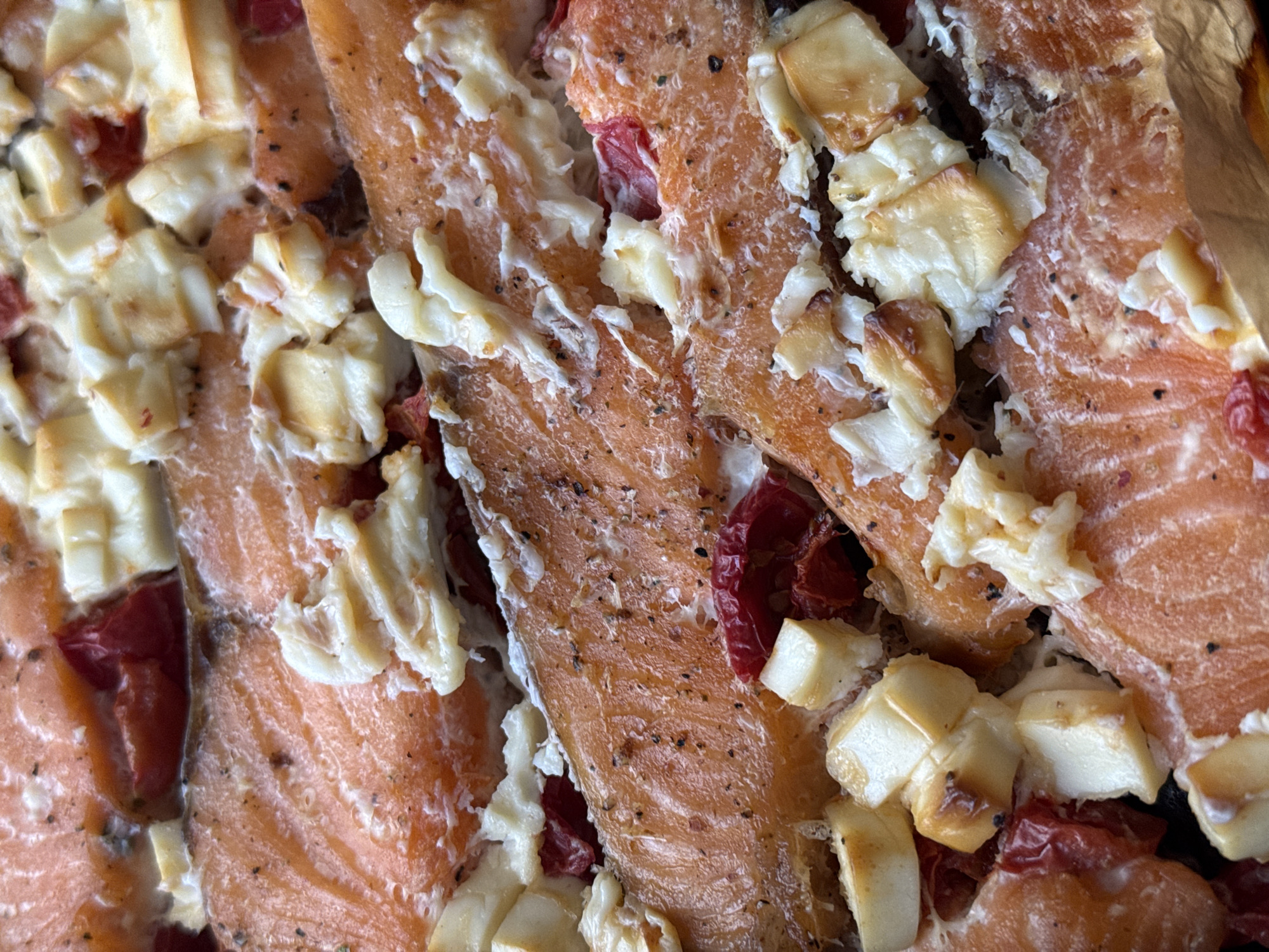 A close-up, overhead shot displays several cooked salmon fillets generously topped with white cheese and red diced vegetables. The salmon has a vibrant orange-pink color, typical of cooked fish, and shows visible flaky textures. Dark specks, likely ground pepper or other seasonings, are visible across the surface of the fillets. Irregularly shaped chunks of white cheese, possibly feta or a similar soft variety, are scattered across the salmon; some pieces show a light golden-brown char, indicating they have been baked or broiled. Small, dark red pieces, resembling chopped sundried tomatoes or roasted red peppers, are interspersed among the other ingredients. The overall scene suggests a freshly prepared meal, likely a main course. The lighting is bright and even, highlighting the details and textures of the food. There are no human subjects, discernible location cues beyond the food itself, or visible text in the image.