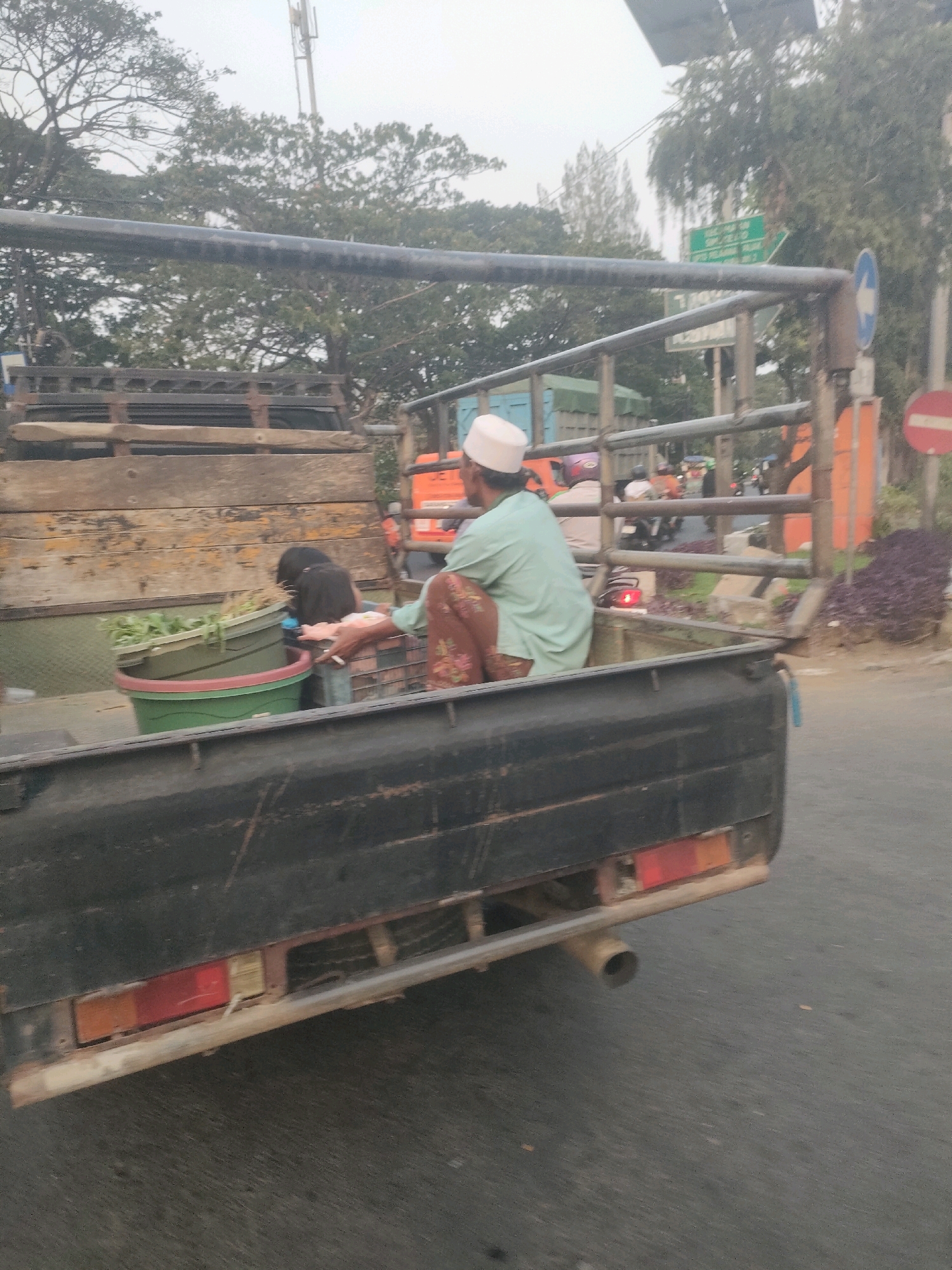 The image captures a rear view of a pickup truck in motion, likely on a street in Surabaya, Indonesia. The truck bed contains several objects and two individuals. A person wearing a light blue long-sleeved shirt and a white cap is seated, facing away from the camera, with their right hand visible. Next to them, a second person, possibly a child, with dark hair, is also seated and partially obscured. Green leafy vegetables are contained within a large green plastic tub and a smaller green bucket, both positioned in the truck bed. The truck's black exterior is weathered, with visible rust and damage. In the background, traffic can be observed, including motorcycles, and urban infrastructure such as trees, a cell tower, and traffic signs, one of which is a circular red and white prohibition sign. The overall scene suggests a typical day on the road in a bustling Indonesian city.