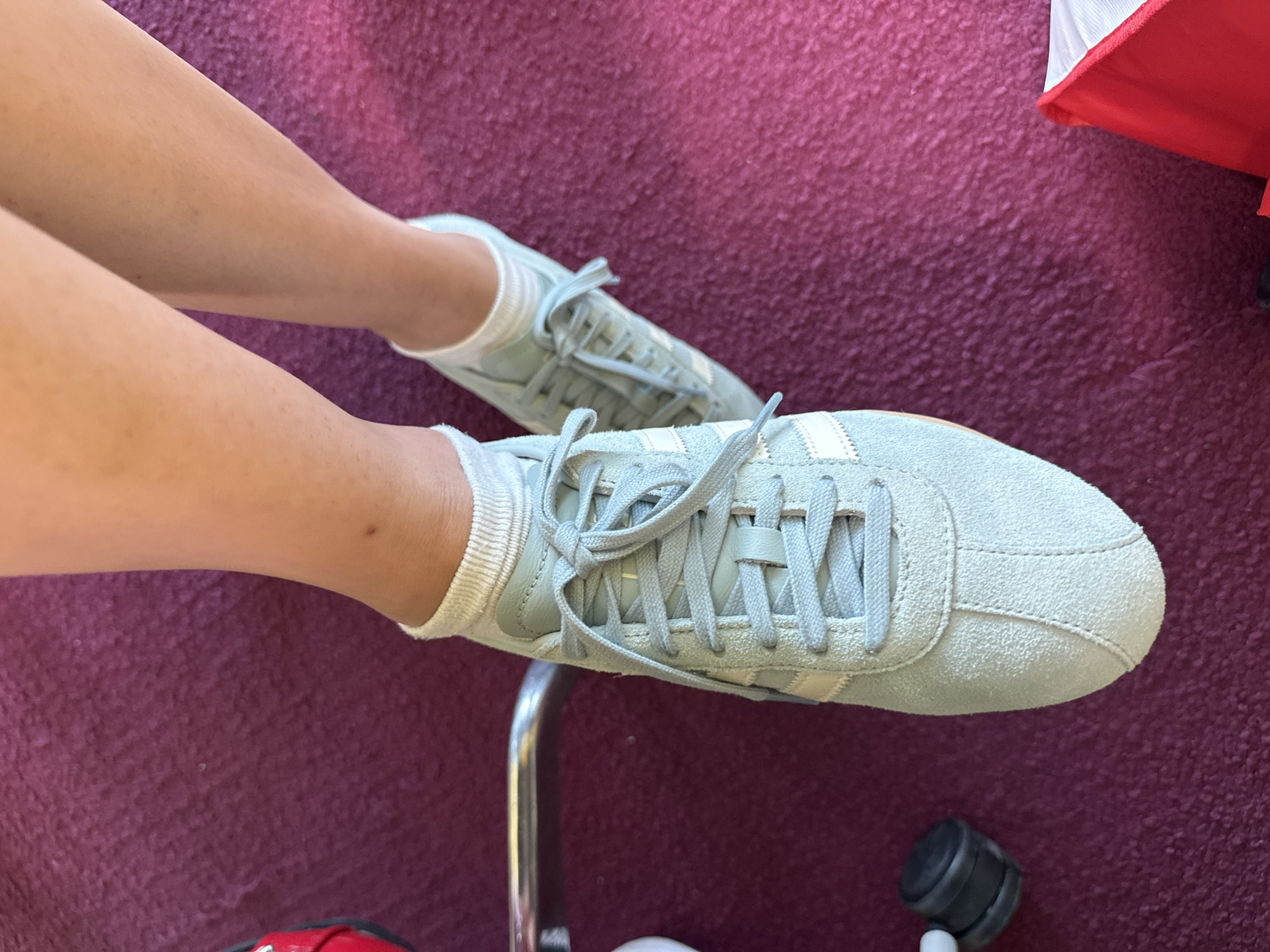 This high-angle, close-up image captures a person's legs and feet resting on a dark red or maroon carpet. The person's legs, which appear tanned, are seen from above, wearing white ankle socks and light blue/grey suede sneakers. The sneakers feature distinct white three-stripe branding on the sides and are laced up with matching blue laces, suggesting a classic athletic style like the Adidas Gazelle.
The feet are positioned casually, with the right foot closer to the foreground and slightly angled, while the left foot recedes into the background. The textured surface of the carpet provides a soft, uniform backdrop to the footwear.
In the lower left, a shiny silver metallic pole is partially visible, hinting at nearby furniture or equipment. Towards the bottom right corner, a small black cylindrical object with a white element is present. A bright red and white object is also partially seen in the upper right corner of the frame. The lighting is bright and even, indicating a daytime setting, likely indoors. The image conveys a sense of comfort and relaxation. No discernible text is visible on any of the subjects. The location context provided is Mexico City, Mexico.