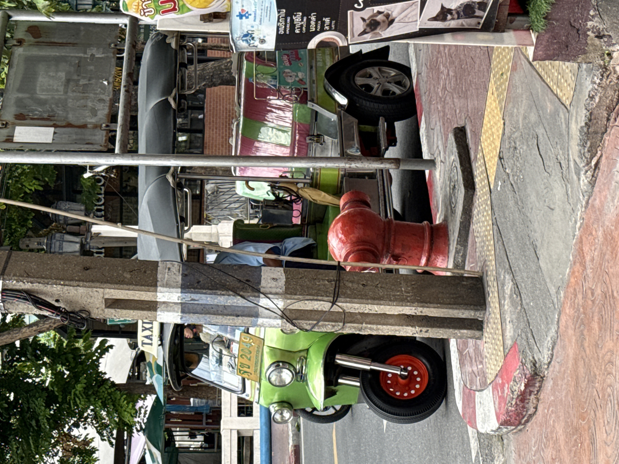 The image depicts a street scene in Bangkok, Thailand, focusing on a bright green tuk-tuk parked near a sidewalk. The tuk-tuk has a "TAXI" sign on its roof and a license plate reading "รบ 2049". The vehicle's side is decorated with green and pink stripes. In the foreground, a red fire hydrant stands on a sidewalk with textured paving stones and a red and white curb. A concrete utility pole with exposed wires is to the left of the fire hydrant. In the background, trees, buildings, and street signs are visible. A person in a blue shirt is seated in the tuk-tuk. The overall impression is of a typical urban environment in Bangkok.