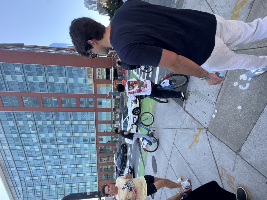 The image depicts an outdoor urban street scene in the Seaport District of Boston, United States. The background is dominated by One Marina Park Drive, a multi-story building featuring a red brick lower section and extensive upper levels constructed with a reflective glass curtain wall.
Five individuals are present in the foreground and midground. In the immediate right foreground, a male subject is viewed from behind, wearing a dark short-sleeved t-shirt, light-colored trousers, and dark sunglasses. He holds a small multi-colored object in his right hand. To his left, a partial view of another individual's lower legs and feet on the sidewalk is visible. Further left, a male subject wearing a yellow short-sleeved t-shirt with a graphic and black shorts is shown in motion, suggestive of jogging, with earphones in both ears. Two other male subjects are riding blue and white Bluebikes rental bicycles; one wears a dark t-shirt and light jeans, and the other wears a white graphic t-shirt with a print on the back and dark pants. Both cyclists are positioned on a green-painted bike lane adjacent to the concrete sidewalk.
The street behind the cyclists contains vehicular traffic, including a white sedan and a dark SUV. The concrete sidewalk features expansion joints and indistinct spray paint markings in yellow and white. Two metal utility access panels are embedded in the sidewalk. The sky above the buildings is clear blue.