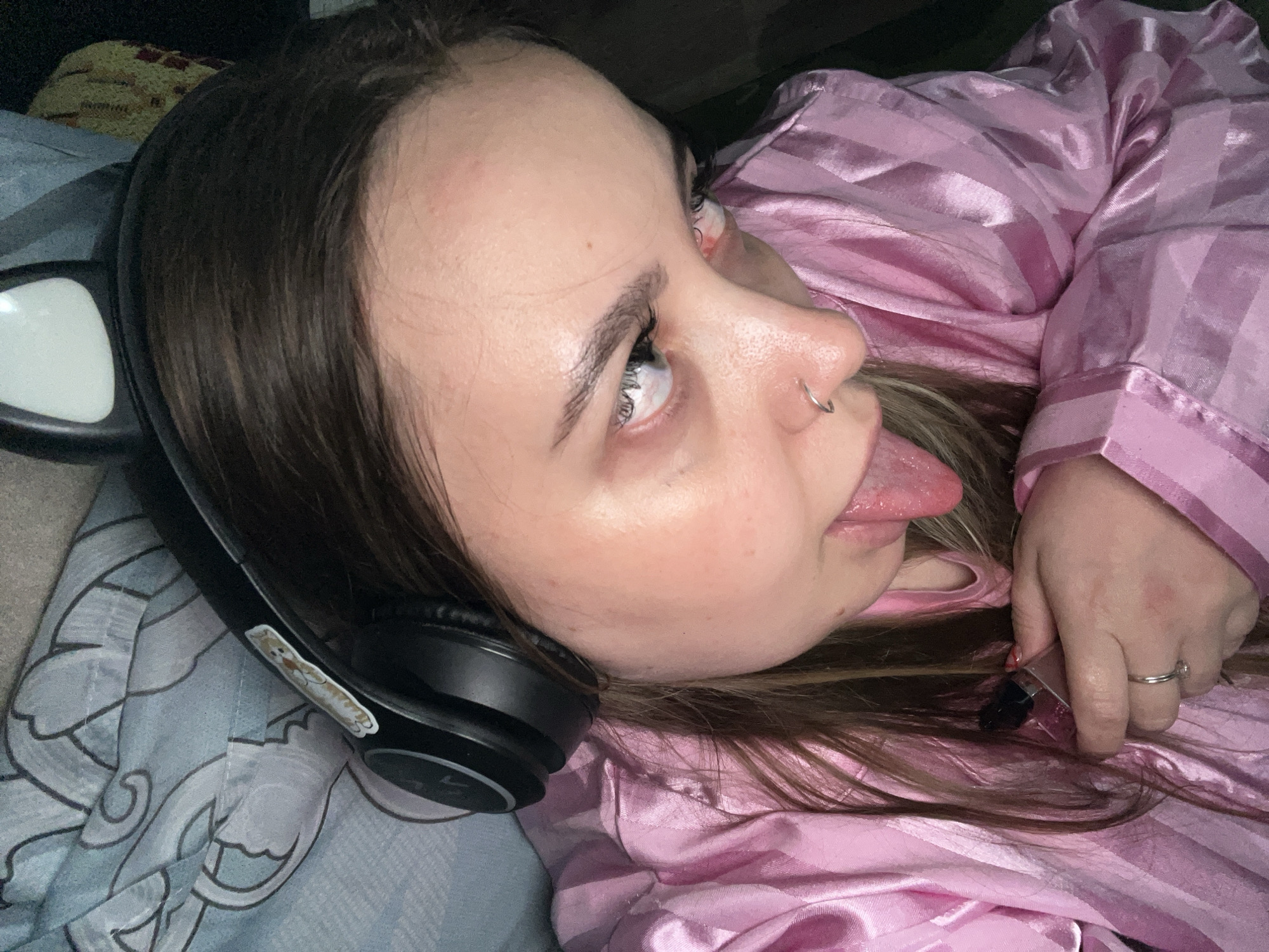 A young woman is lying down, wearing headphones with a sticker on them and a pink striped pajama top. She has a nose ring and is sticking her tongue out. Her hair is brown with lighter streaks. She is holding a small, possibly cosmetic, item in her hand. She appears to be lying on a patterned blue and white surface, likely a bed. There is no indication of Novopokrovskaya, Russia, in the image.