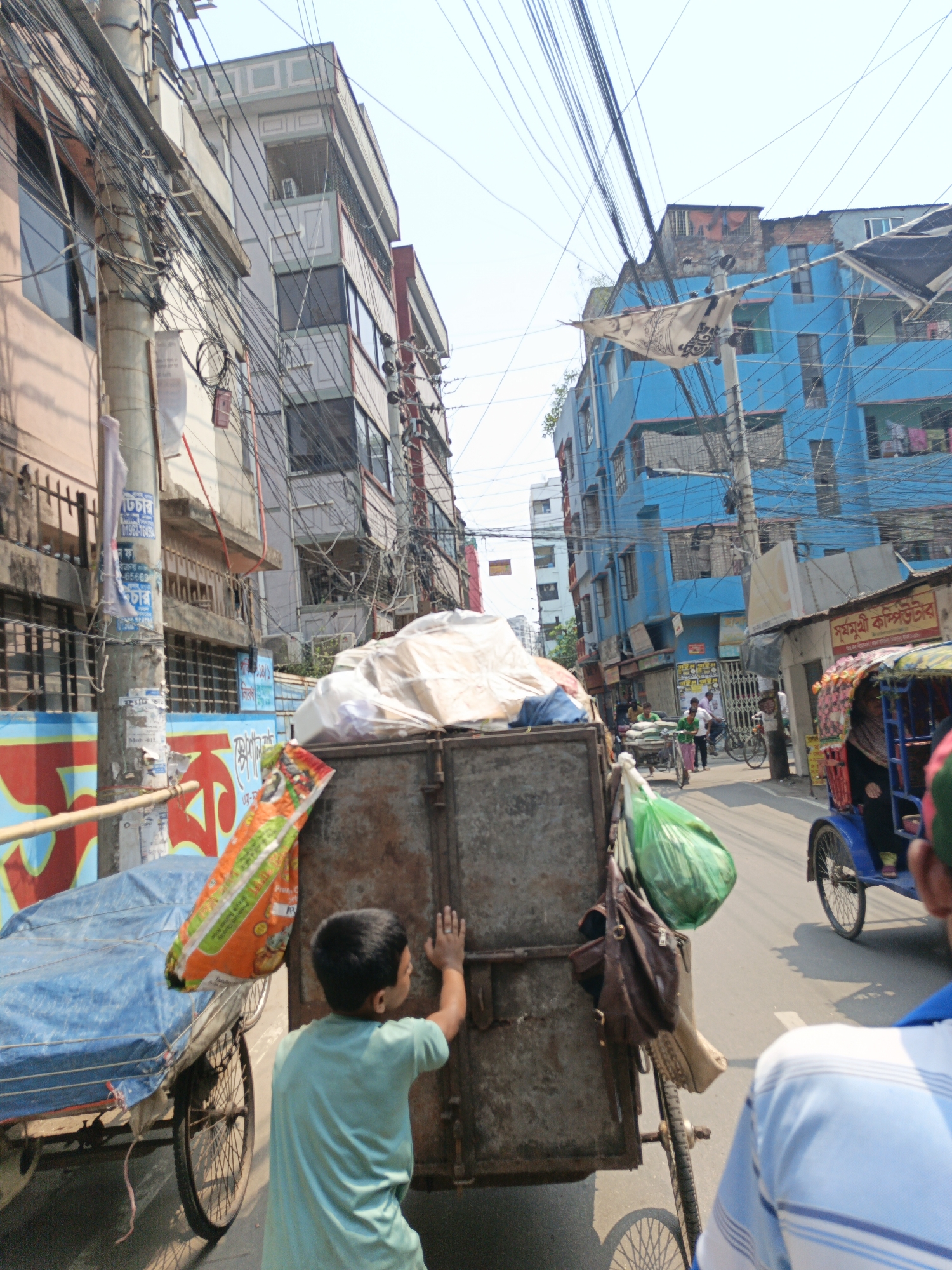 A street scene in Dhaka, Bangladesh. In the foreground, a young boy wearing a turquoise shirt is pushing a large, rectangular, metal cart filled with white and tan fabric, and an orange bag is attached to the side. To the left, there's a cart covered with a blue tarp, and behind the metal cart, there's a green plastic bag and a brown purse. A blue rickshaw with a passenger is visible on the right side. Two adults are in the bottom right corner, wearing shirts with blue stripes.
In the background, a blue building dominates the right side, while other buildings in varying colors line the narrow street, and an array of electrical wires crisscrosses the sky. People are walking and cycling in the street. The buildings and wires are signs of a densely populated urban area.