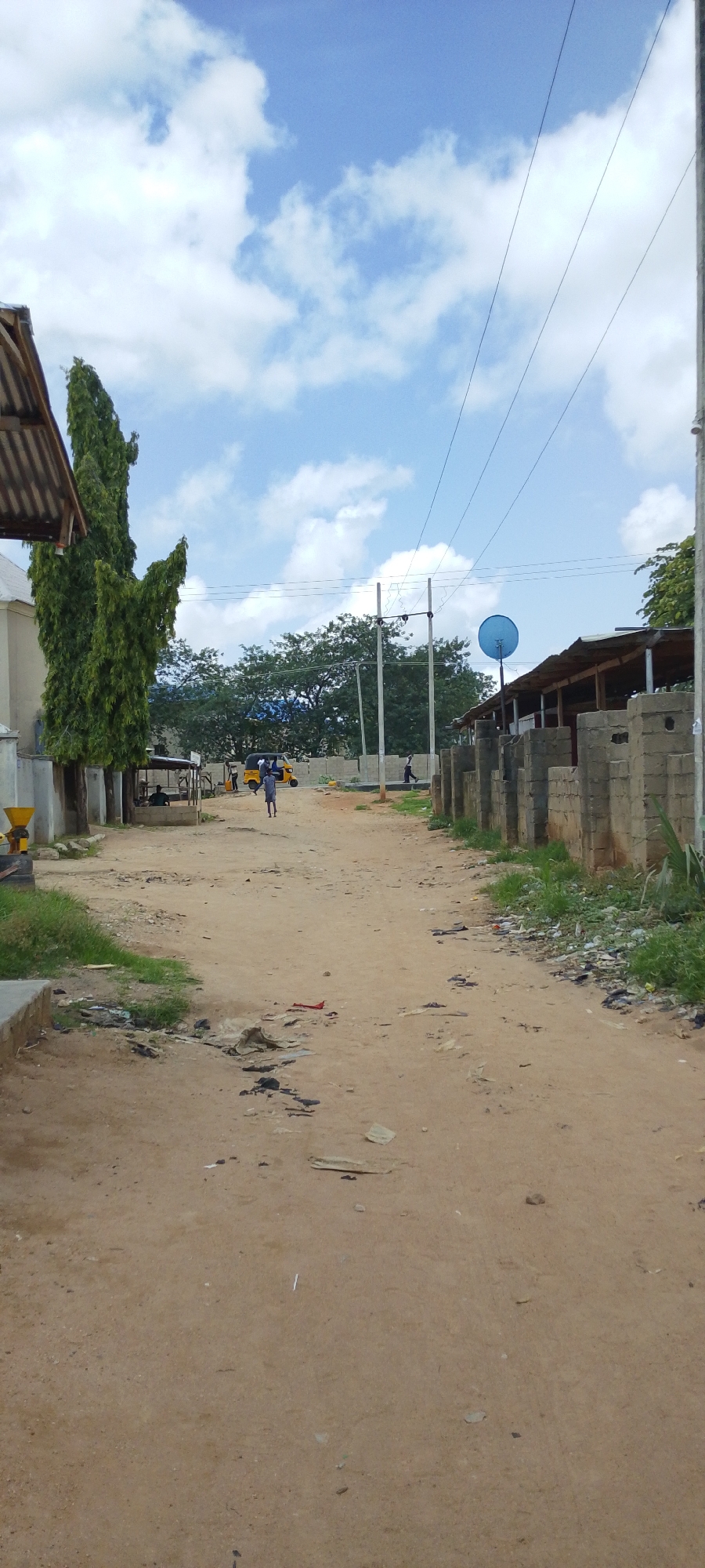 The media file depicts an outdoor scene of a dirt road in what appears to be a village or rural area. The road is unpaved and shows signs of wear and tear, with some debris scattered on the ground. On the left side of the road, there is a tall, slender green tree and a portion of a building with a corrugated roof. Further down the road, a yellow auto-rickshaw is visible, with a few people gathered around it. On the right side of the road, a low wall constructed of concrete blocks runs along the path, with a covered structure built alongside it, possibly a shop or shelter. A blue satellite dish is mounted on a pole behind this structure. In the background, there are more trees, and electrical wires are strung across the sky, supported by utility poles. The sky is blue with scattered white clouds, suggesting it is daytime with fair weather. A few individuals are walking or standing in the distance, indicating some level of activity. The overall impression is a casual, everyday scene in a developing or rural environment. There is no visible text in the image.