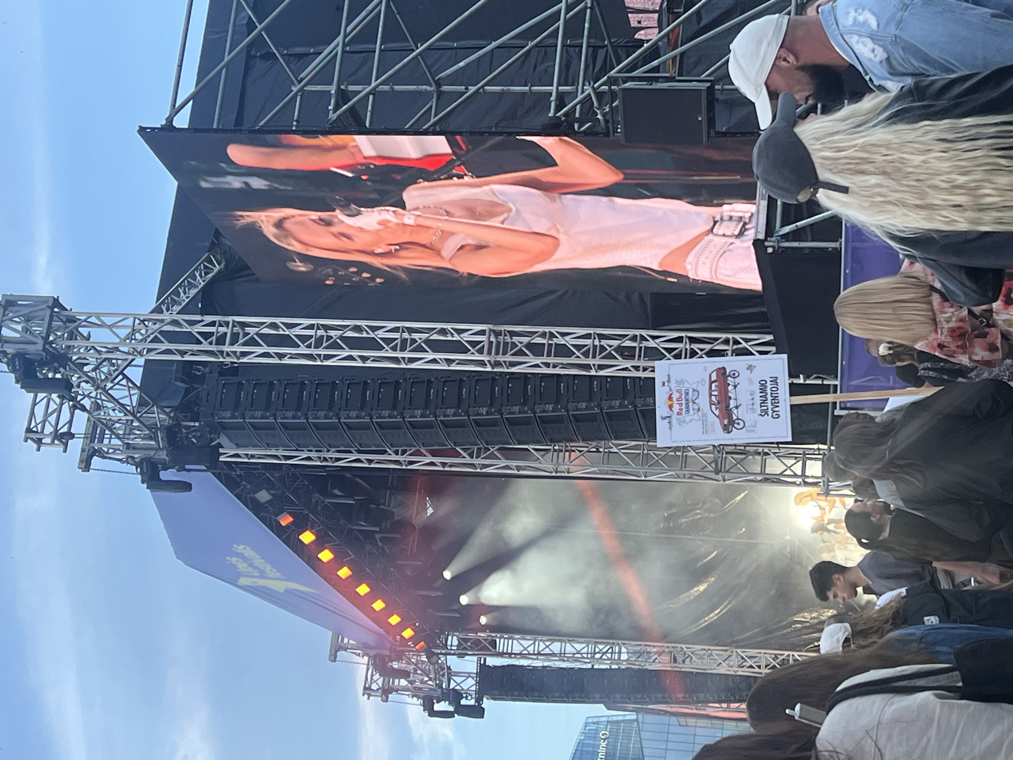 The image depicts an outdoor festival stage with a large LED screen displaying a blonde woman singing. The stage is equipped with lighting and sound equipment. In the foreground, a crowd of people is visible, watching the performance. A sign with a Red Bull logo and the text "ŠILTNAMIO GYVENTOJAI" is also present. The background shows modern buildings, suggesting an urban setting. The event appears to be "Upės Festivalis" in Vilnius, Lithuania, indicated by the banner on the left side of the stage.