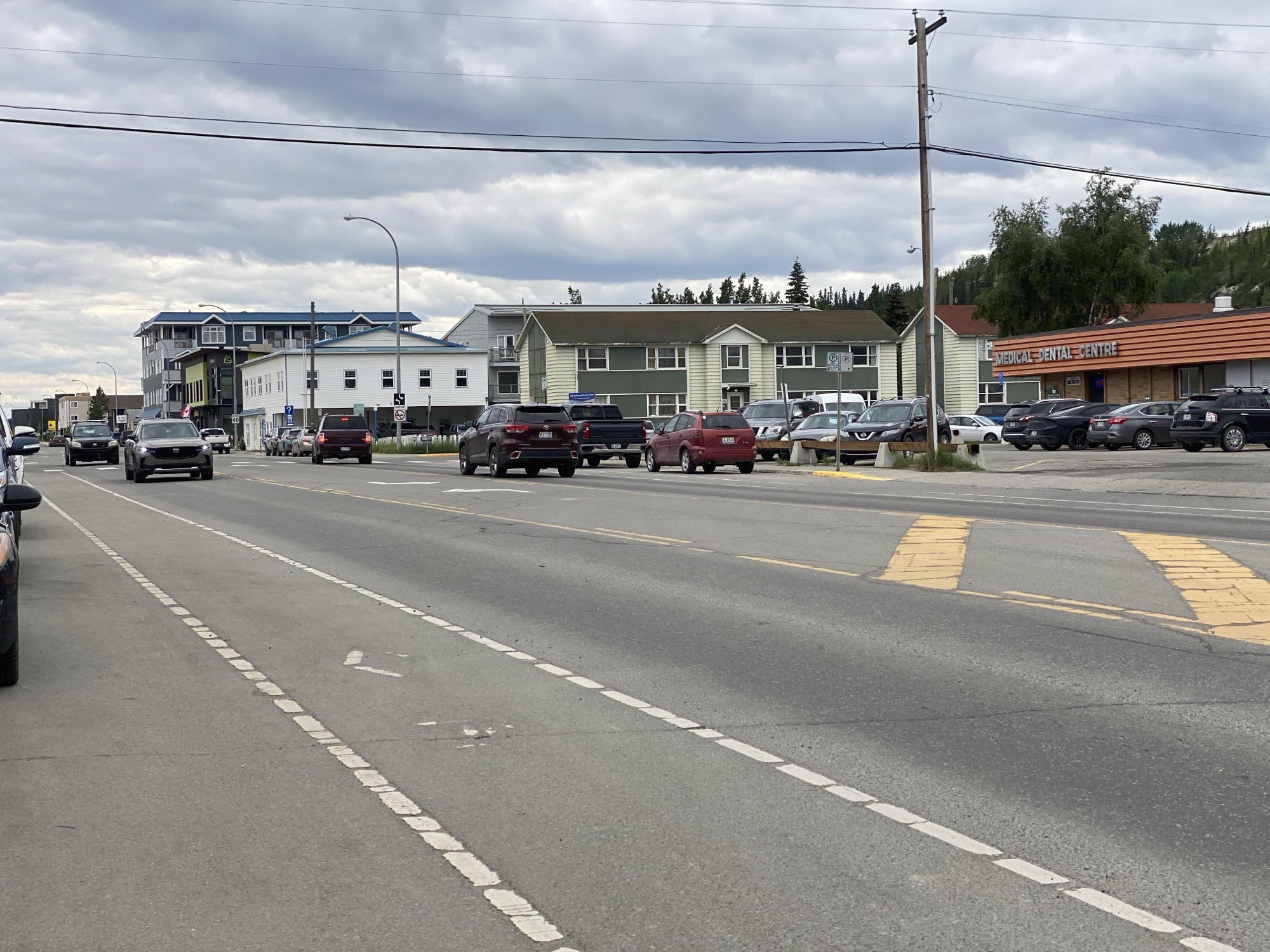 The image captures a street scene in Whitehorse, Canada, specifically looking east on Fourth Avenue near the intersection with Alexander Street. The weather appears overcast. On the left side of the road, several vehicles are parked or moving, including a black SUV, a dark sedan, and a red SUV. The road itself is asphalt with clear lane markings, including a dashed white line separating lanes and a solid yellow line on the far side.
Buildings line the north side of the street. The most prominent building in the foreground on the right is the Medical Dental Centre, an orange-paneled building with signage. Adjacent to it, to the west, is a pale green building with white trim, possibly an office or residential complex. Further west are multi-story buildings, one with a blue roof and modern architecture, and another smaller white building.
The background shows a densely forested hillside, characteristic of Whitehorse's natural surroundings. A wooden utility pole with electrical wires is visible on the right side of the image, and a street lamp stands near the center of the frame. The overall impression is of a commercial and residential area within a northern city.