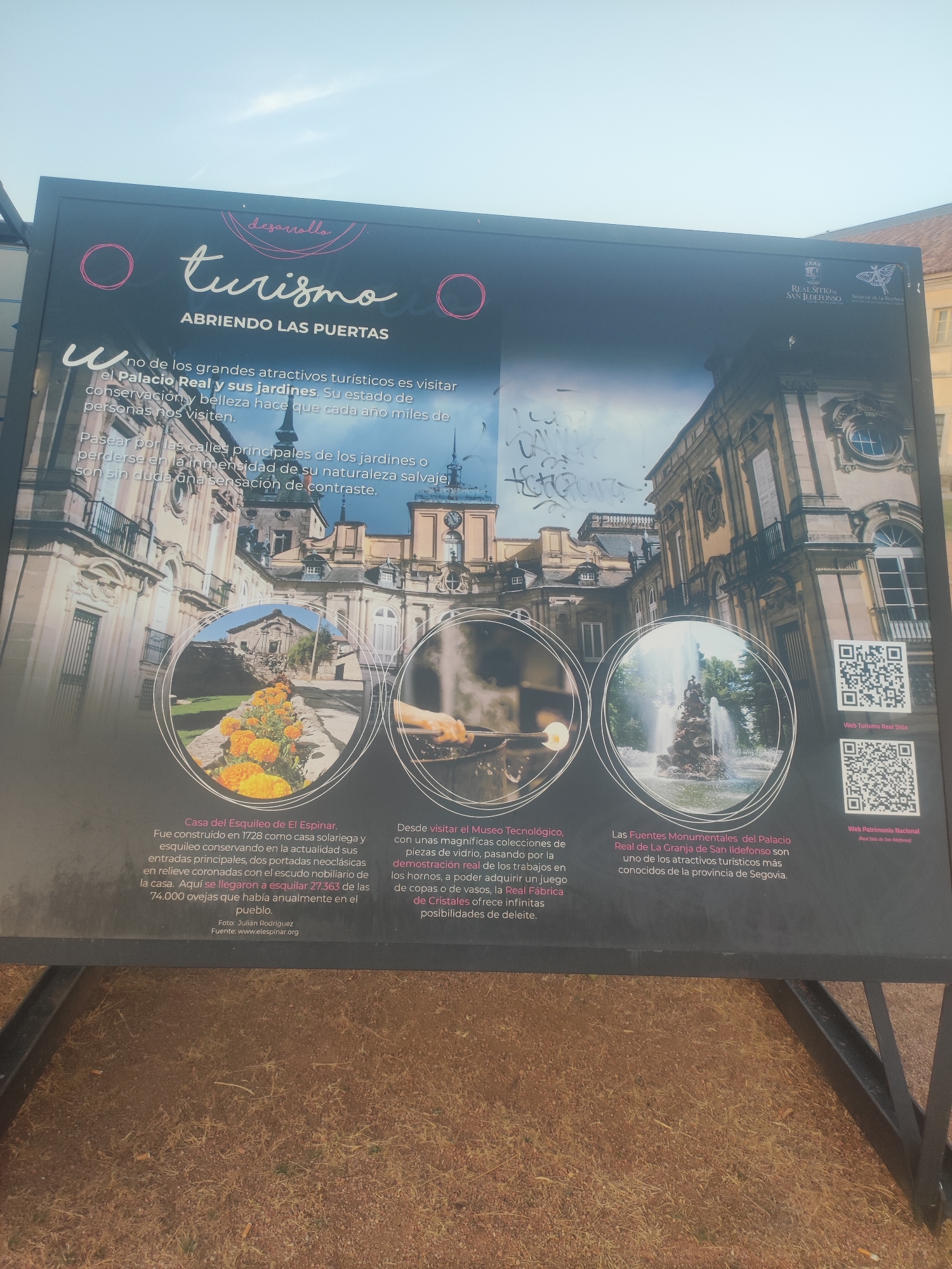 A sign is displayed outdoors, likely in Real Sitio de San Ildefonso, Spain, promoting tourism. The sign features a large, dark background with text and images. The title "desarrollo turismo" is prominently displayed in stylized white and pink lettering, followed by "ABRIENDO LAS PUERTAS".
The main image on the sign is a composite of the Royal Palace of La Granja de San Ildefonso and its gardens, with three circular insets. The left inset shows a close-up of bright orange flowers. The middle inset depicts a hand working with molten glass, suggesting a craft or museum exhibit. The right inset showcases a grand fountain with water jets.
Below the insets, descriptive text is provided in Spanish, detailing attractions such as "Casa del Esquileo de El Espinar," "Museo Tecnológico," and "Las Fuentes Monumentales del Palacio Real de La Granja de San Ildefonso." QR codes and website addresses are also visible on the right side of the sign.
The sign is mounted on a black metal frame, and the ground in front of it is covered with dry grass and dirt. The sky above is overcast and pale. There are no people or animals directly visible on the sign itself. The overall impression is an informational and promotional display for a tourist destination.