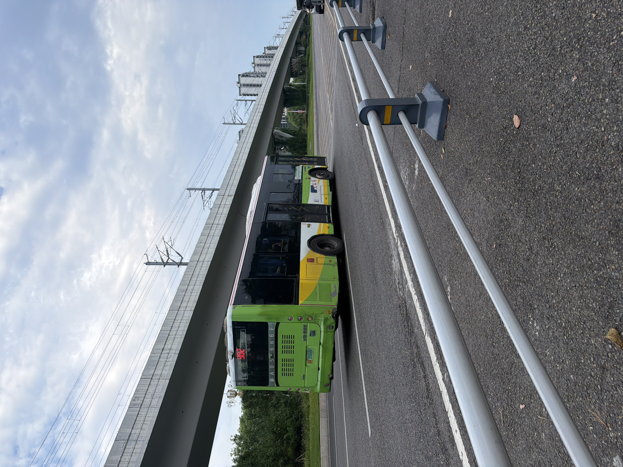 A green and white public bus with the number "302" displayed on its front is driving on a paved road. The bus has several windows and a license plate with the characters "A 05666F". In the background, there is a large elevated structure that appears to be an overpass or highway, with electrical wires running overhead. Beyond the overpass, there are several modern buildings and trees. The sky is cloudy. The road is marked with white lines and there is a metal barrier along the right side of the road, with grey posts and silver rails. The scene appears to be in an urban or suburban area.