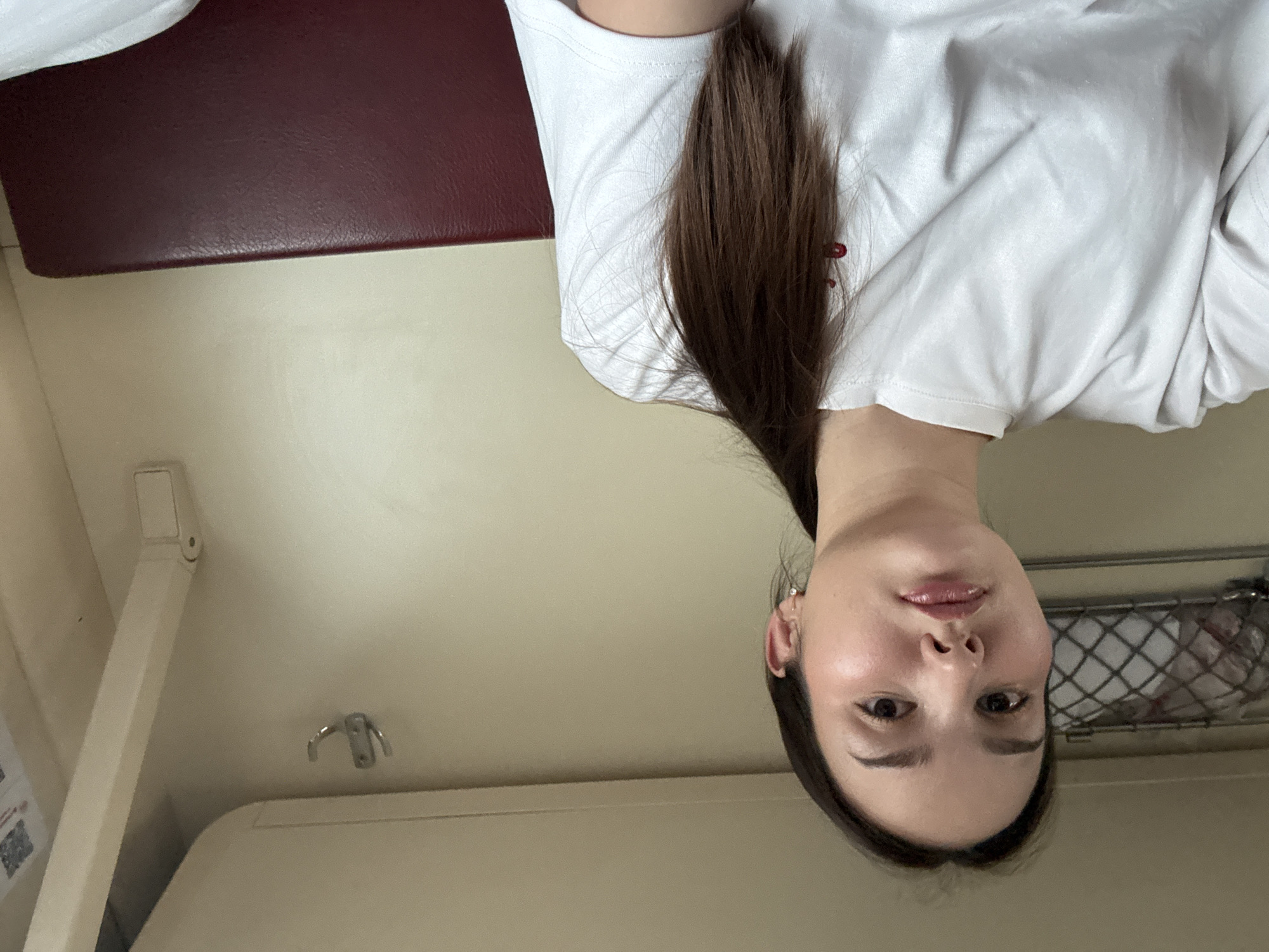 The image depicts a female individual within what appears to be a train compartment, consistent with typical Russian railway carriages, presumably en route to or from the city of Cheremkhovo, Russia.
**People:** One female, appearing to be a young adult with fair skin, dark brown hair styled in a ponytail, dark eyes, and full lips. She wears a plain light-colored (possibly white) crew-neck t-shirt with a small red detail on the left chest, and small stud earrings are visible in both ears. She is looking directly at the camera with a neutral expression.
**Objects and Environment:** The individual occupies the left and central foreground of the frame. The background reveals the interior of a confined space. To the upper left, a metal mesh luggage rack is present, containing a white, possibly textile, item. A silver-colored double coat hook is affixed to the light-colored wall paneling on the upper right. Below and to the right of the individual, a section of a dark red (maroon) upholstered seat or bunk is visible. The walls and ceiling are uniformly light-colored, consistent with interior paneling of a transport vehicle. A light-colored horizontal trim piece runs across the upper wall/ceiling junction, and a vertical structural element is present on the far right. Indiscernible blurred text or a graphic is partially visible on the far right wall panel. The overall lighting is even and suggests an interior, possibly artificial, light source.