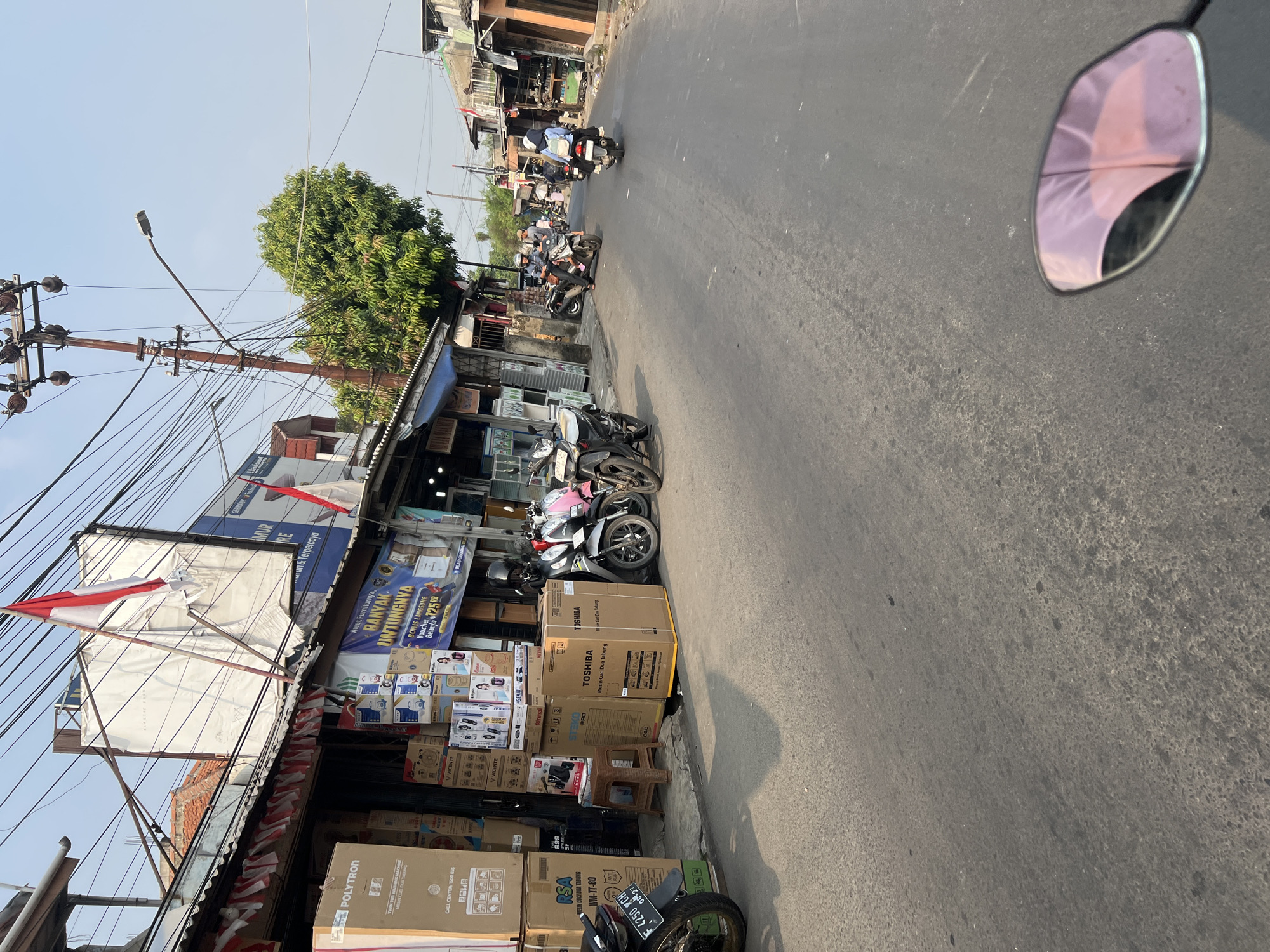 The image depicts a street scene in Citeureup, Indonesia, characterized by a row of shops selling appliances and electronics, as indicated by stacked boxes from brands like Polytron and Toshiba. Several motorcycles are present, with some parked and others being ridden by individuals. One person on a motorcycle is visible in the foreground, and others are further down the street. The shops have awnings and banners, with one banner displaying "BANYAK ANTIHUTIINNYA," suggesting a sale or promotion. Indonesian flags are displayed, one prominently on a pole above the shops and smaller ones decorating the storefronts. Electrical wires and a utility pole are visible overhead. The street is paved, and the environment appears to be an urban or semi-urban commercial area. The lighting suggests it is daytime, likely morning or afternoon.