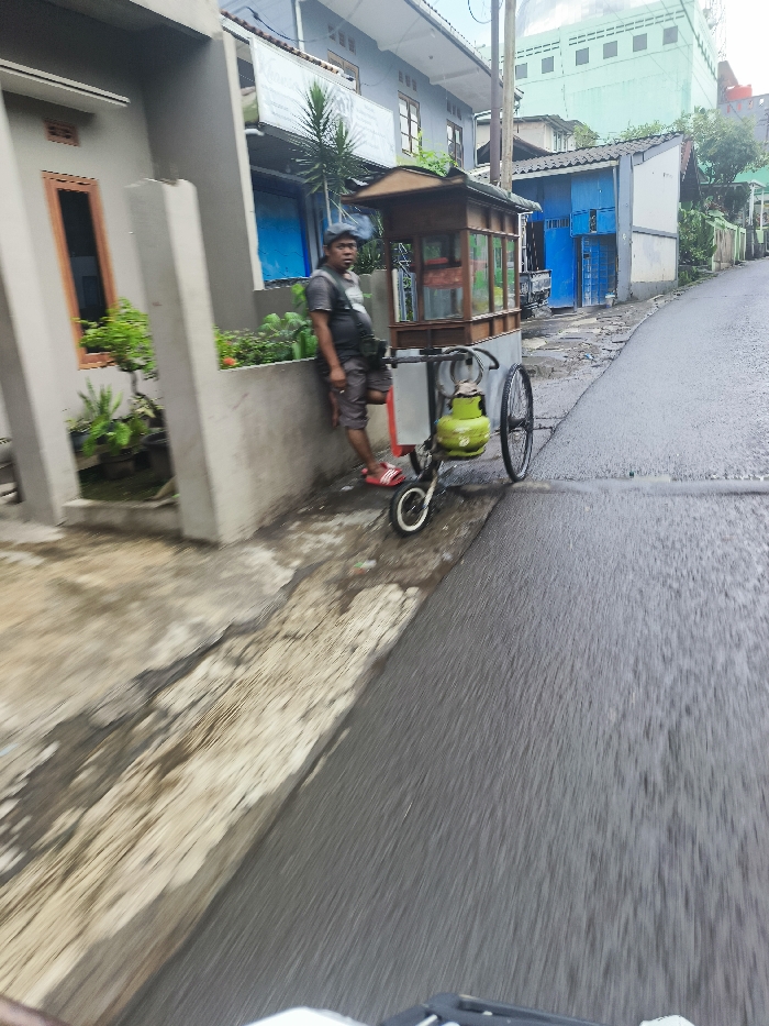 The image shows a man standing next to a small, wooden food cart with a single wheel and a green gas cylinder attached. The cart is parked on the side of a paved street in Cimenyan, Indonesia. The man is wearing a grey shirt, blue jeans, and a dark baseball cap. The street is wet and the buildings lining the street are residential, with a blue door visible. The man appears to be waiting for customers. The camera is located on a moving vehicle, with the street in the foreground. The image is slightly blurred due to the movement of the camera.