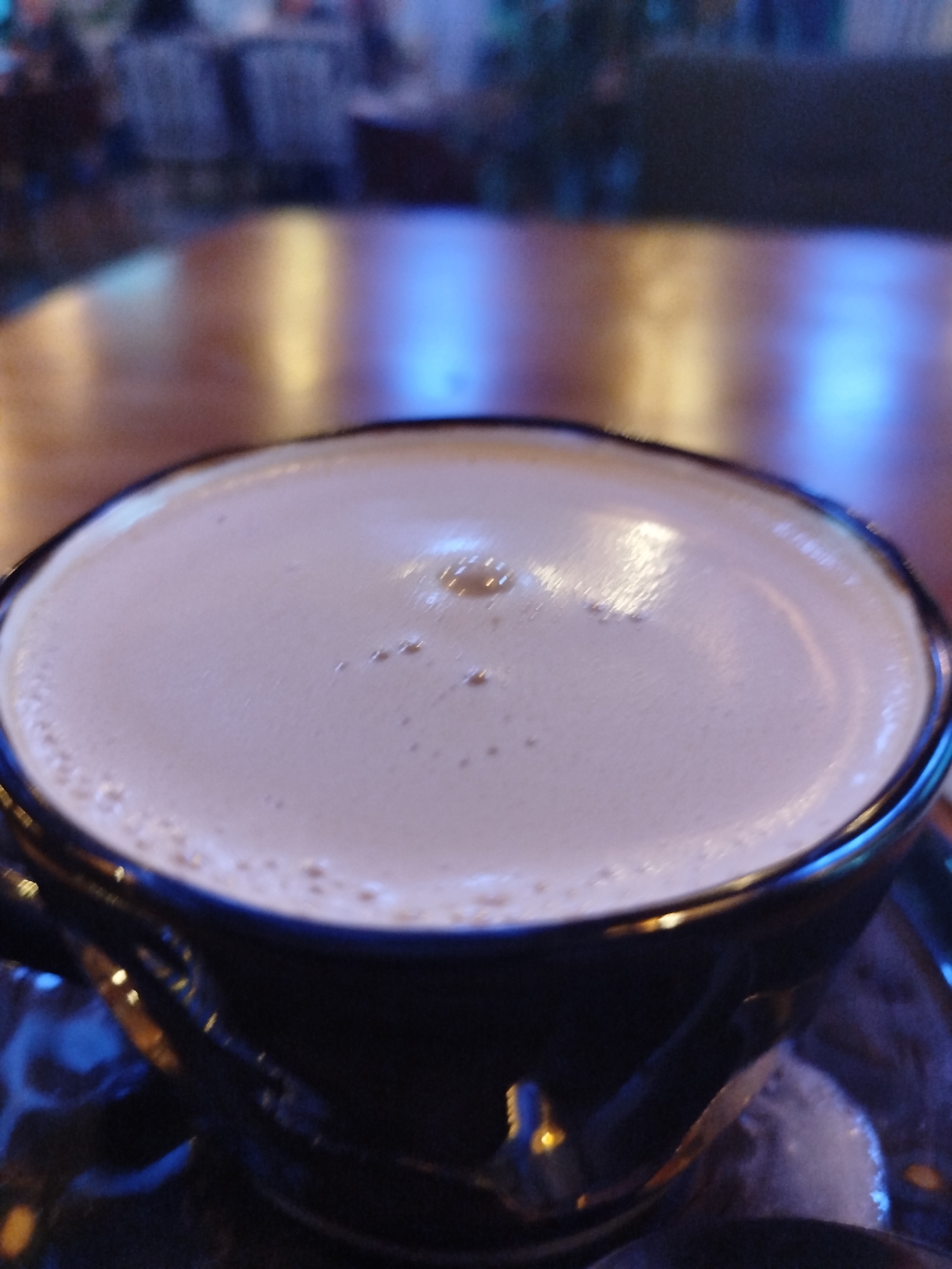 A close-up, blurry shot shows the top of a frothy beverage in a dark mug, placed on a wooden table. The background is out of focus, with hints of blue and yellow lights suggesting an indoor setting with ambient lighting, possibly a cafe or restaurant. No specific landmarks or locations in Kremenchuk, Ukraine, are identifiable.