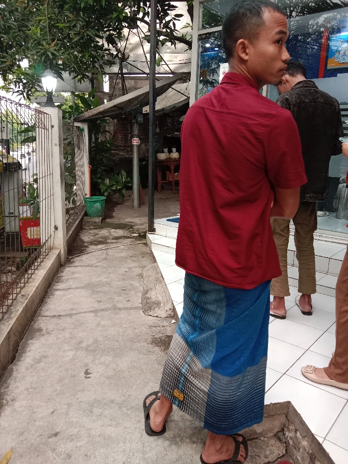 The scene takes place in the Kecamatan Ngaliyan district of Semarang, Indonesia, at the coordinates -6.992 latitude and 110.353 longitude. A man in a red shirt and blue sarong is standing on a concrete sidewalk, facing the camera. His left foot is on the ground, and his right foot is raised as if he is about to take a step. He has short dark hair and his expression is neutral. Behind him, to the right, is a second man wearing a blue denim shirt and brown pants. He is looking to his right and standing with his weight shifted to his left foot. In the background, a third person, whose legs can be seen, is standing on a tiled floor, looking down towards the sidewalk. The sidewalk is lined with a metal fence on the left side of the frame. The concrete surface of the sidewalk is cracked and uneven. The area behind the men is partially obscured by a leafy tree and the side of a building. A small green trash can sits on the sidewalk near the fence. The tiled floor to the right of the sidewalk appears to be part of the entrance to a building. There is an out-of-focus image of a car and what appears to be a street in the distance. The day is partly cloudy and the lighting is bright.