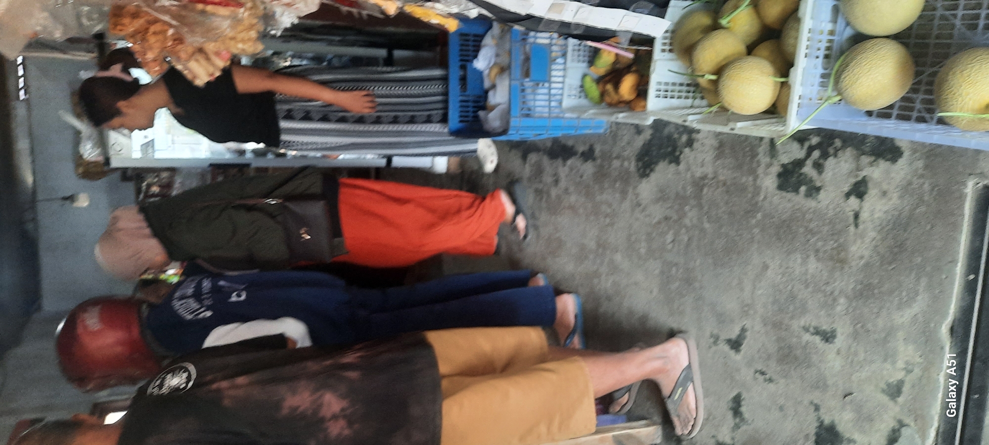 In Selopuro, Indonesia, a group of people are gathered near a stall or shop. In the foreground, a person wearing tan shorts and grey flip-flops stands with their left foot partially visible. To their right, another individual wears blue pants and appears to be wearing blue flip-flops. A person in a dark top with a patterned tie-dye effect and tan shorts is also visible on the far left.
Behind them, a person wearing a red helmet and a blue jacket with white accents is seen. Next to them, a person in a light pink hijab and a dark green jacket is visible, holding a black purse. Further back, a person in an orange skirt and dark sandals is standing. To the right, a woman with her hair pulled back in a ponytail wears a black t-shirt and a black and white patterned skirt.
Several crates are present. A blue crate in the middle ground contains some items wrapped in plastic. To the right, a white crate holds mangoes, and below it, another white crate is filled with melons. The ground appears to be concrete with some dark stains. The overall lighting suggests it is daytime. The "Galaxy A51" watermark is visible in the bottom left corner.