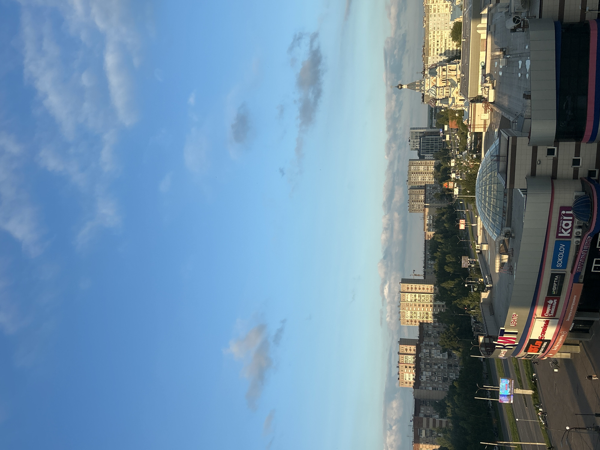 This is a vertical, eye-level, medium shot of a city scene. The sky, filled with scattered clouds, occupies the left two-thirds of the frame. The right third of the image shows a cityscape with multi-story buildings. In the foreground on the right, a modern shopping center with a glass dome is visible, featuring colorful signage with Russian text such as "ЦЕНТР," "SOKOLOV," and "Kai." Other buildings of various architectural styles, including one with onion domes, are scattered throughout the urban landscape. Trees line the streets and surround some of the buildings, suggesting a park or green spaces within the city. The lighting indicates it is either sunrise or sunset, casting long shadows and bathing the scene in a warm glow. The image appears to be taken from an elevated position, overlooking the city. No people are clearly discernible in the frame. The overall impression is one of a bustling urban environment during a tranquil time of day.