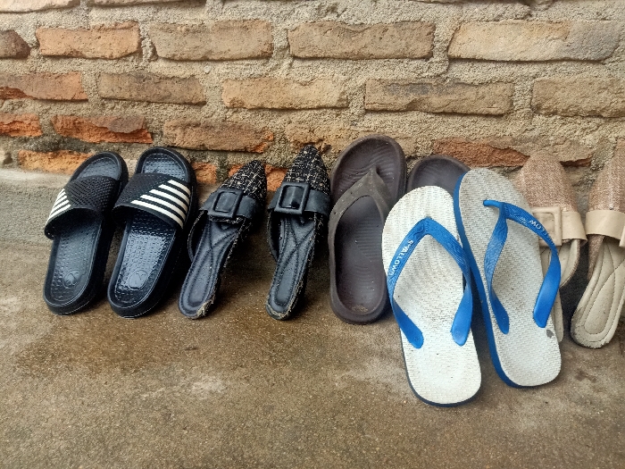 The image captures a collection of seven pairs of footwear arranged neatly against a weathered brick wall on a damp concrete surface.

On the far left are two pairs of black slides. The first pair has a textured black strap, while the second features a black strap adorned with three white stripes. Both black slides show an embossed "Y" logo on their inner soles. Adjacent to these are two black low-heeled slippers or mules, characterized by a textured, possibly woven, upper material and a prominent rectangular buckle on the strap.

Following these, a pair of dark brown flip-flops is visible. To their right, a pair of white flip-flops with vibrant blue straps stands out. The brand name "SWALLOW" is clearly legible in white text on the blue straps of these flip-flops. The last pair on the far right consists of light brown or beige slip-on shoes, each featuring a square buckle on the top.

The background is dominated by an old brick wall, showing variations in brick color from reddish-brown to lighter tones, with some signs of age and discoloration. The foreground is a grey concrete or paved ground that appears wet or damp, indicating recent moisture, possibly from rain or cleaning. The lighting suggests it is daytime, using natural light. No individuals are present, and no particular activity is taking place; the shoes are simply laid out. The scene depicts a casual outdoor setting in Pubian, Indonesia.