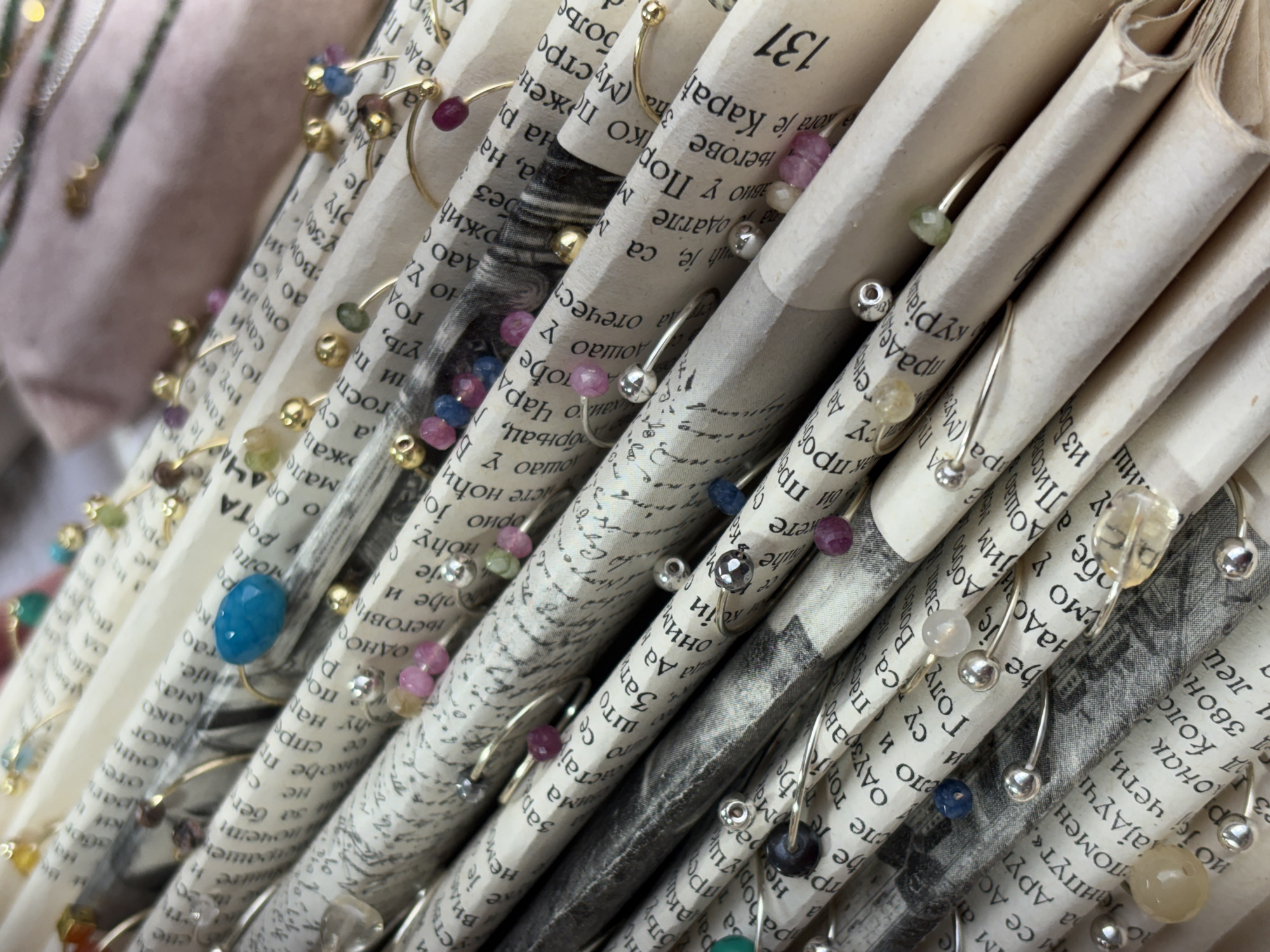 The image shows a close-up of rolled-up pages from a text, likely from a book or newspaper, with numerous silver or gold-colored wire rings attached. Each ring is adorned with a small, colorful bead or stone. The text is printed in a Cyrillic alphabet, possibly Serbian, and the number "131" is visible on one of the pages. The focus is on the texture of the paper, the variety of colored beads, and the placement of the rings. The background is out of focus, suggesting the subject is isolated for artistic or commercial purposes. The image doesn't suggest anything about the location of Belgrade, Serbia, in particular.