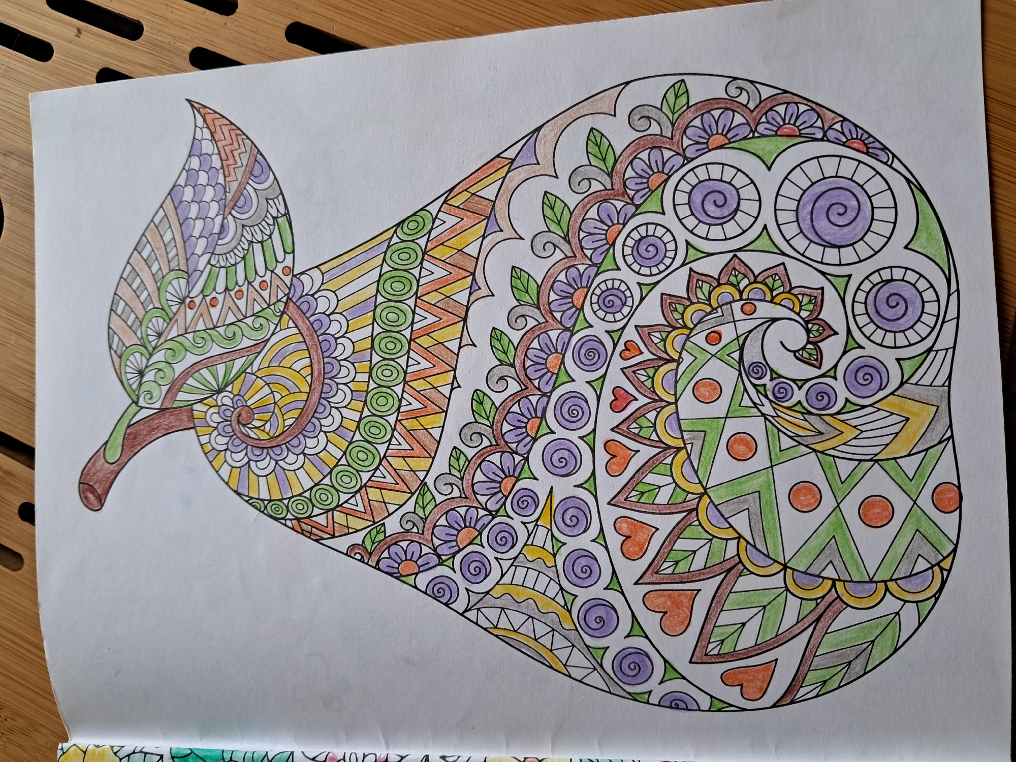 This is a close-up, eye-level, slightly angled shot of an adult coloring book page featuring a zentangle-style pear. The pear is intricately detailed with various geometric patterns and decorative elements. The coloring is incomplete, with many areas left blank, but some sections are colored in shades of green, yellow, orange, purple, and brown. The pear is positioned centrally on the white page. To the left of the coloring page, a sliver of the adjacent page with more coloring book designs is visible, featuring a border of colorful abstract patterns. The coloring book is lying open on a light brown wooden surface with a visible slatted texture, suggesting a table or desk. The lighting is natural, and the overall impression is of a creative activity in progress.