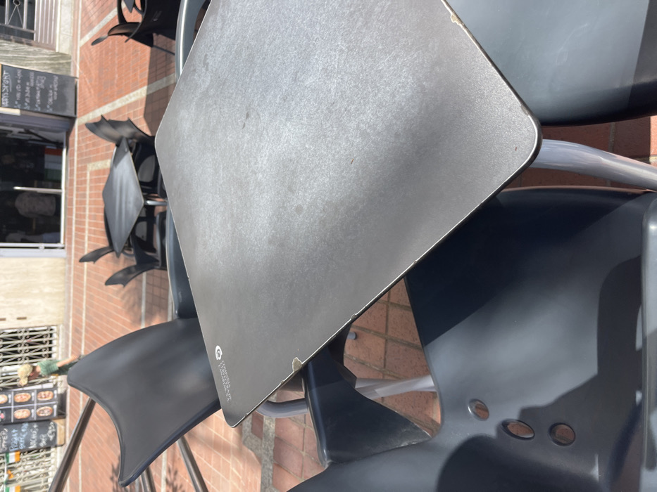 The image shows a close-up view of a square, metal table with a smooth, grey surface, located on a brick-paved patio, in the heart of Las Palmas de Gran Canaria, Spain, at the given coordinates. The table is surrounded by black plastic chairs. Three of the chairs are empty, while the fourth is obscured by the table's surface. The table is in the center of the frame and is partially covered in shadow. The table's surface features a light, faded inscription, possibly a brand or logo. The patio is visible in the background, showing other tables and chairs, along with a building wall, and the faint outline of a figure walking away.