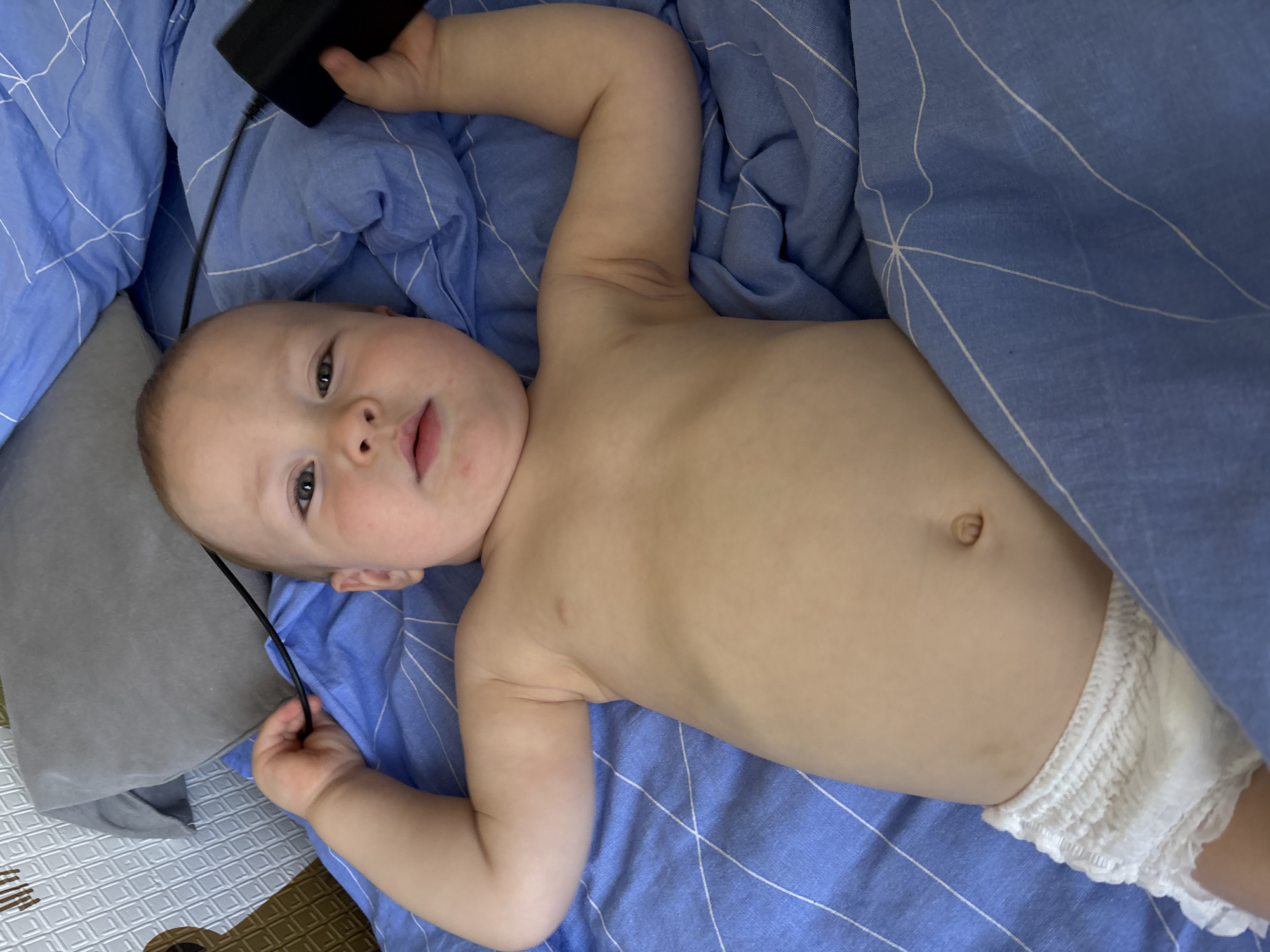 A close-up, overhead shot shows a baby lying on their back on a blue, striped blanket. The baby is wearing a white diaper with ruffled edges. The baby has fair skin, light brown hair, and is looking up with wide, curious eyes. Their left arm is extended upwards and is holding a black object, possibly a charger or a small electronic device, connected to a black cable. The baby's face is slightly turned to the left, and their mouth is open, suggesting they might be about to speak or have just spoken. The lighting appears to be indoor, suggesting it is daytime. The background consists of blue bedding and a gray pillow. The overall mood is one of peacefulness and innocent exploration.
