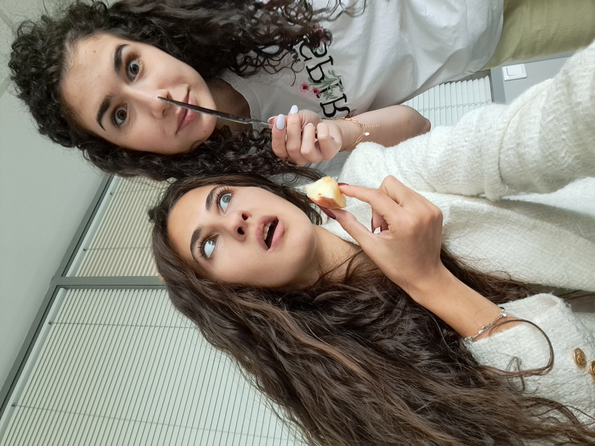 Two women are captured in a selfie-style image. The woman in the upper portion of the frame holds a butter knife up to her nose. She has curly dark hair, fair skin, and dark eyebrows. She is wearing a white t-shirt with floral decorations and writing. Her fingernails are painted white. She wears a gold bracelet on her left wrist.
The woman below her is looking upward, with her mouth slightly open, holding a piece of fruit. She has long wavy brown hair, fair skin, and dark eyebrows. She is wearing a white sweater with gold buttons. She wears a silver bracelet on her left wrist.
The background features a window with horizontal blinds and a wall painted a light color.
There is no indication in this image of the location being in the city of Sofia, Bulgaria.