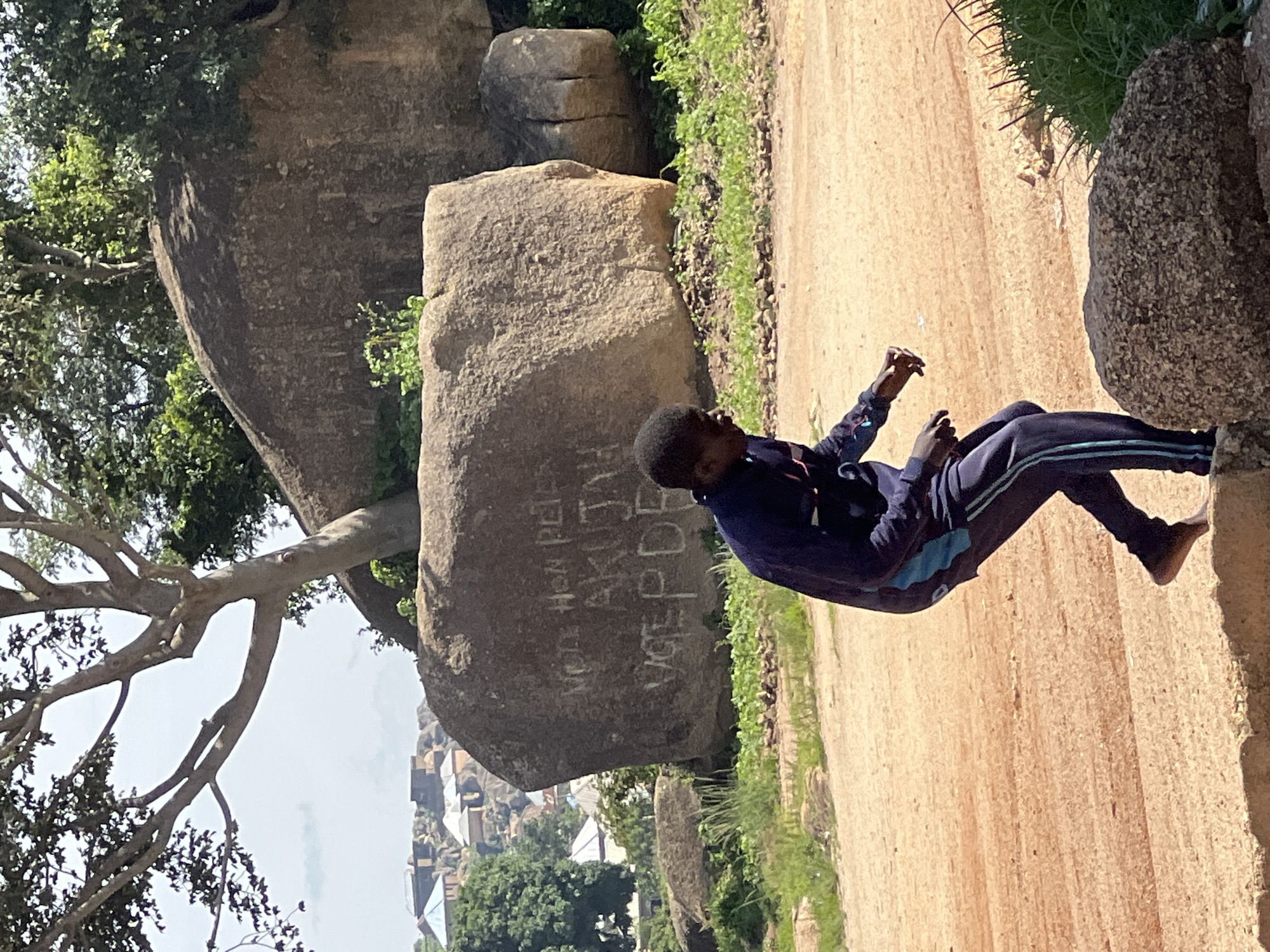 In the city of Jos, Nigeria, a young Black male is captured in motion, his body turned slightly to the left, one leg bent and his arms extended forward. He wears a dark blue tracksuit with lighter blue accents. The background features large, weathered boulders, one of which is marked with graffiti. The graffiti on the largest boulder appears to read "Vote Hon Peter Akujah Vote PDP". Surrounding the boulders are sparse green vegetation and dry, reddish-brown earth, indicative of the dry season. In the distance, a cluster of buildings with varied roofing materials, including blue and white, suggests a settlement nestled among rocky outcrops. The sky above is a pale, overcast blue.