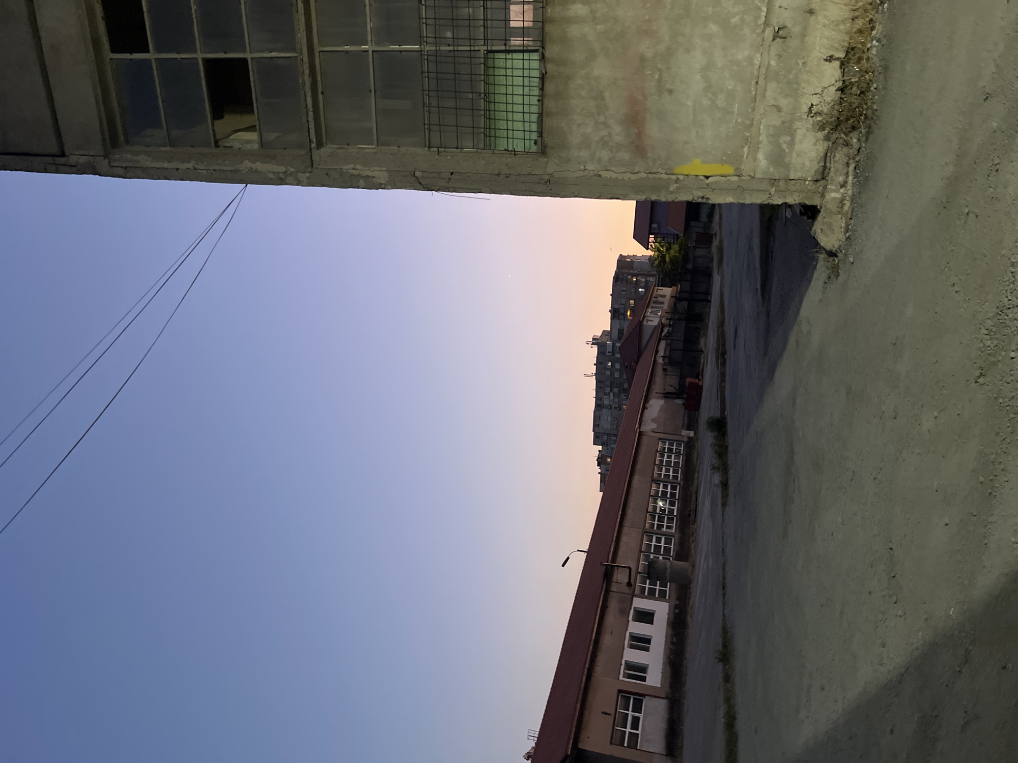 This is a vertical, eye-level, outdoor, nighttime photograph taken in the city of Montana, Bulgaria. The scene depicts an industrial area with a large, dilapidated concrete building in the foreground on the right. This building has multiple windows, some of which are broken or boarded up, and a metal grate on one window. The sky is a gradient of blue transitioning to a pale orange near the horizon. In the background, a long, single-story building with a red roof and numerous windows is visible. Further in the distance, there are apartment buildings. Overhead, power lines are strung across the frame. The ground is a dirt and gravel surface, with shadows cast from the structures. There are no people or animals visible in the image. The overall impression is one of an abandoned or neglected industrial site at dusk.