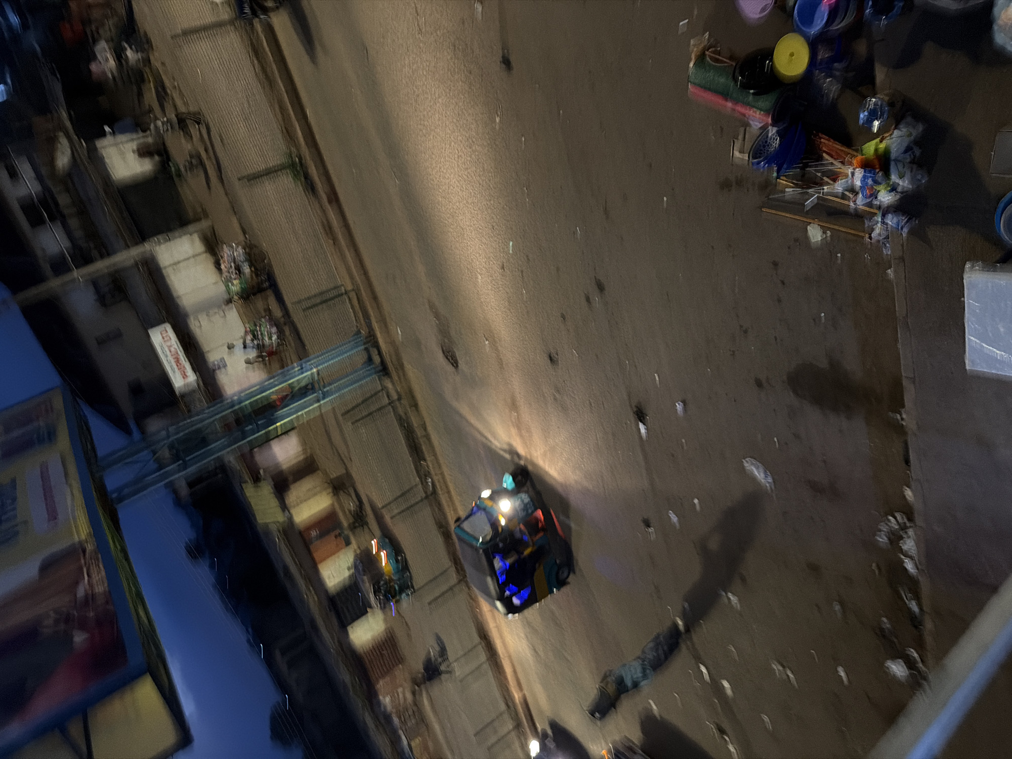 The image depicts a street scene in Bukuru, Nigeria, at dusk. A three-wheeled auto-rickshaw, illuminated by its headlights and interior lights, is the central focus in the foreground, driving on a paved road. The rickshaw has blue and green bodywork with colorful lights. In the background, a pharmacy sign is visible on a building. Several people are walking along the sidewalk and a cart is being pushed. The street appears to be somewhat cluttered with debris. To the right, various household items like buckets, brooms, and mats are displayed for sale. The overall lighting suggests the transition from daylight to night, with artificial lights from buildings and vehicles becoming prominent. The image is slightly blurred, possibly due to movement.