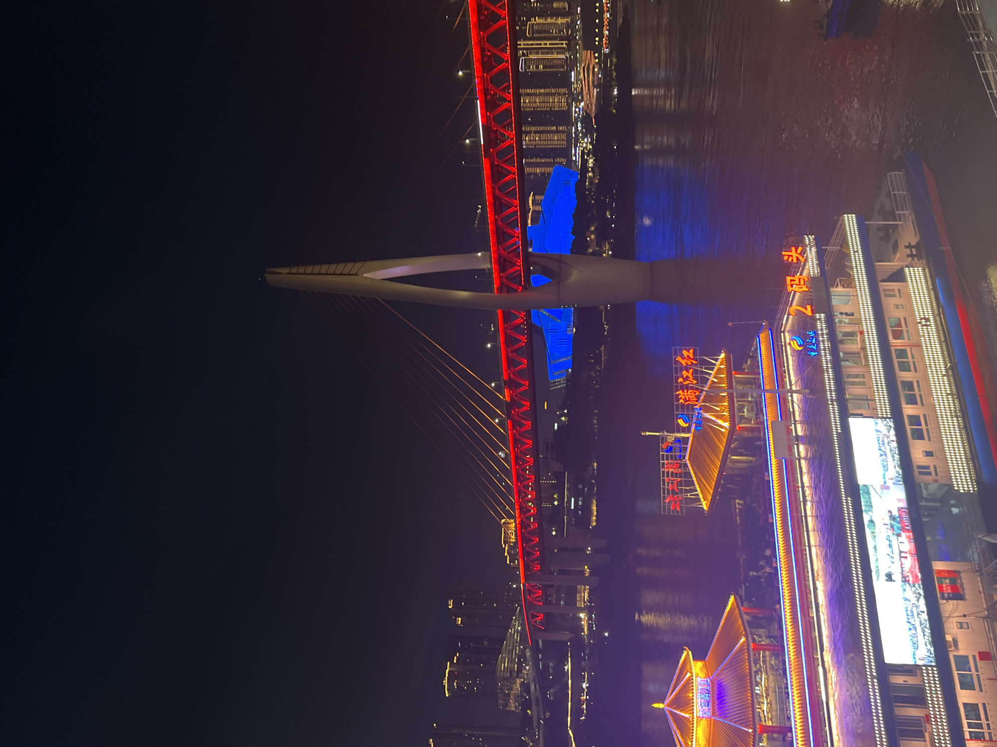 The image shows a nighttime view of the Yangtze River in Yuzhong District, China, with the Hongyadong Bridge illuminated in red and blue lights. A large cable-stayed bridge spans the river, with its white towers and red illuminated deck creating a striking contrast against the dark sky. The water reflects the vibrant lights of the bridge and the city skyline. In the foreground, a multi-story ferry boat is docked, also adorned with colorful lights. The ferry has a large LED screen displaying an image. Two traditional pagoda-style structures with illuminated roofs are also visible on the riverbank, near the ferry. The city skyline in the background features numerous high-rise buildings illuminated with yellow lights, indicating a bustling urban environment. The overall scene is one of urban nighttime splendor, dominated by artificial illumination.