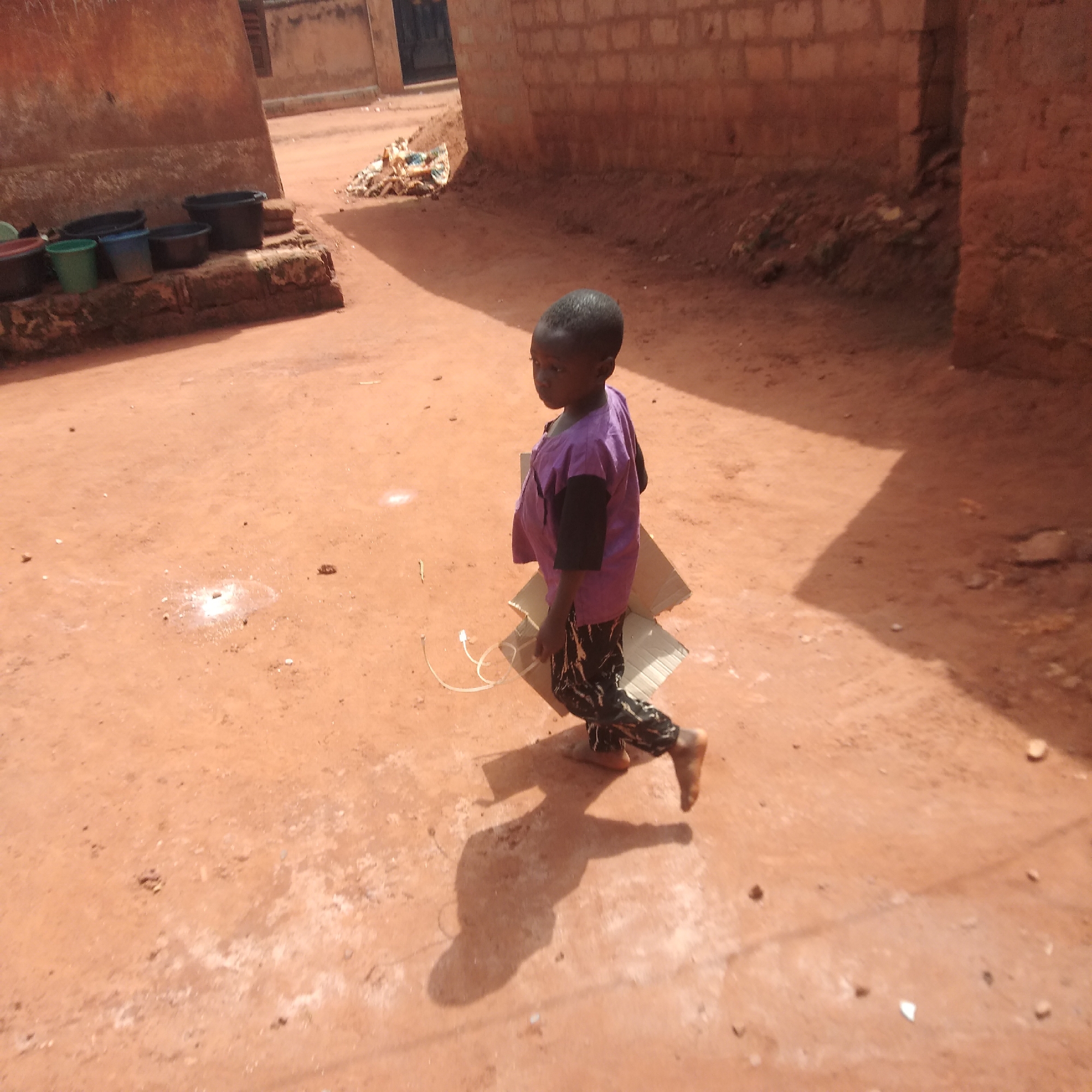 A young child is walking barefoot on a dirt road. The child is wearing a purple shirt, black sleeves, and black patterned pants. They are carrying cardboard boxes with a white cord. To the left, there are containers on a brick structure. In the background, buildings are visible. The location appears to be a rural area, but the exact city is unknown.