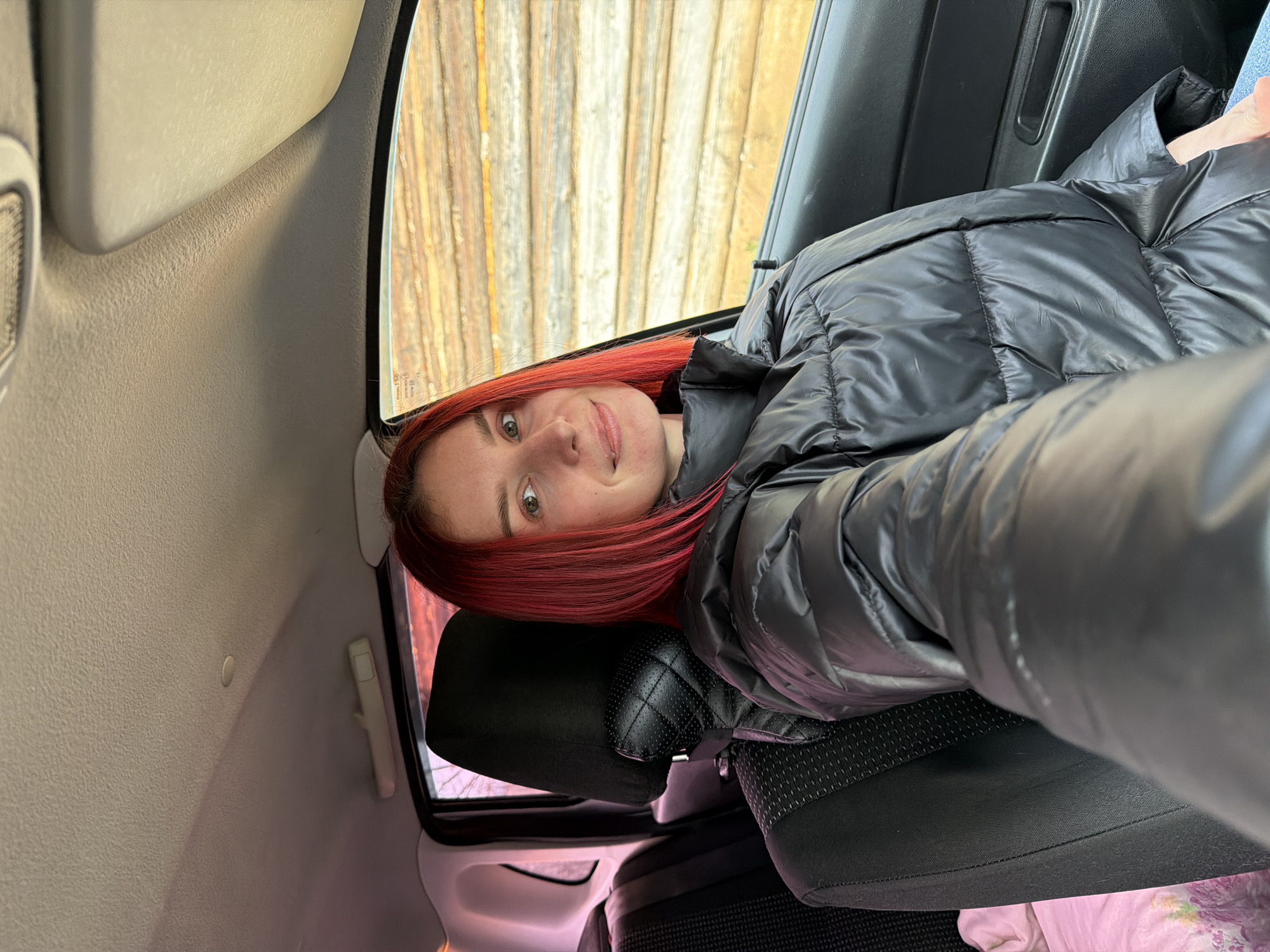 A woman with red hair wearing a black puffer jacket sits in the back seat of a car. Outside the car window, vertical wooden boards are visible. The location is Заболотово, Russia.