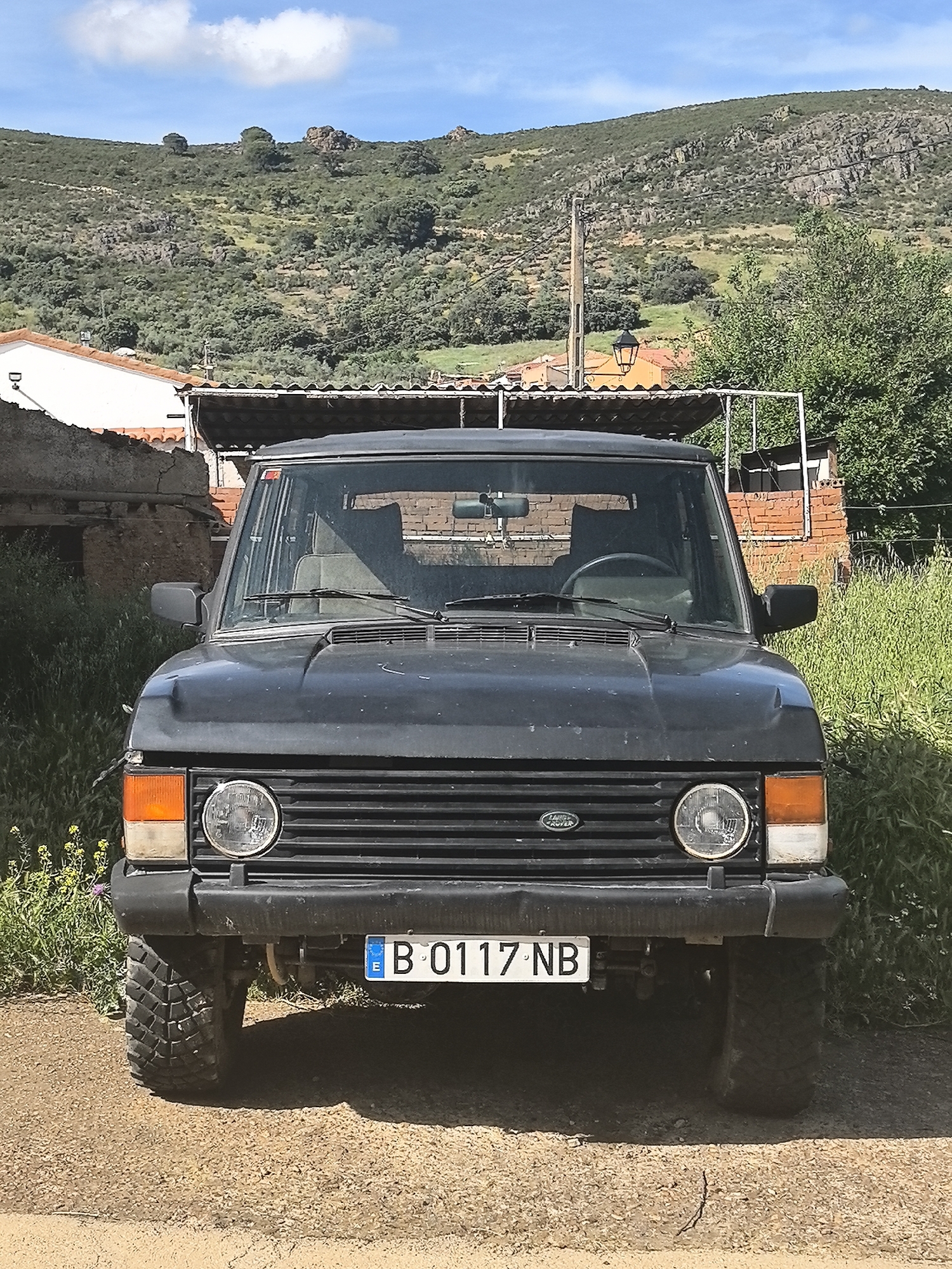 A dark-colored, older model Land Rover is parked on a dirt surface in what appears to be a rural setting. The vehicle faces the camera, showcasing its front grill, headlights, and license plate (B 0117 NB). The background features a landscape with a building, a hillside covered in low vegetation, and a blue sky with clouds. The building appears to be an old structure with a tiled roof. The location is likely Fontanarejo, Spain.
