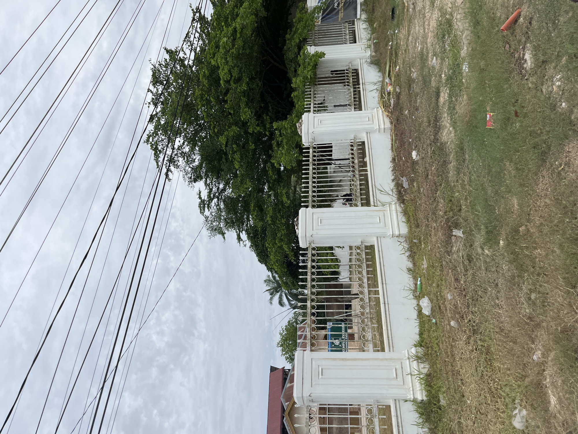 The image captures an outdoor scene in Bundar, Indonesia, featuring a white metal fence bordering a property. Behind the fence, lush green trees and a palm tree are visible, suggesting a tropical environment. The sky is overcast with clouds, indicating it might be a cloudy day. In front of the fence, the ground is covered with dry grass and scattered debris, including plastic bags and a red object that could be a piece of packaging. A portion of a building with a reddish-brown roof is also visible to the left. Several power lines crisscross the sky, dominating the upper left portion of the frame. The overall impression is one of an outdoor, possibly residential or commercial area, with a mix of natural elements and urban infrastructure. No specific activity or event is clearly depicted, and the scene appears static. The weather seems overcast, and the location cues point to a tropical setting. The image is taken from a low angle, looking upwards towards the sky and the fence. Some text is visible on a green sign behind the fence, but it is illegible due to the resolution and angle.